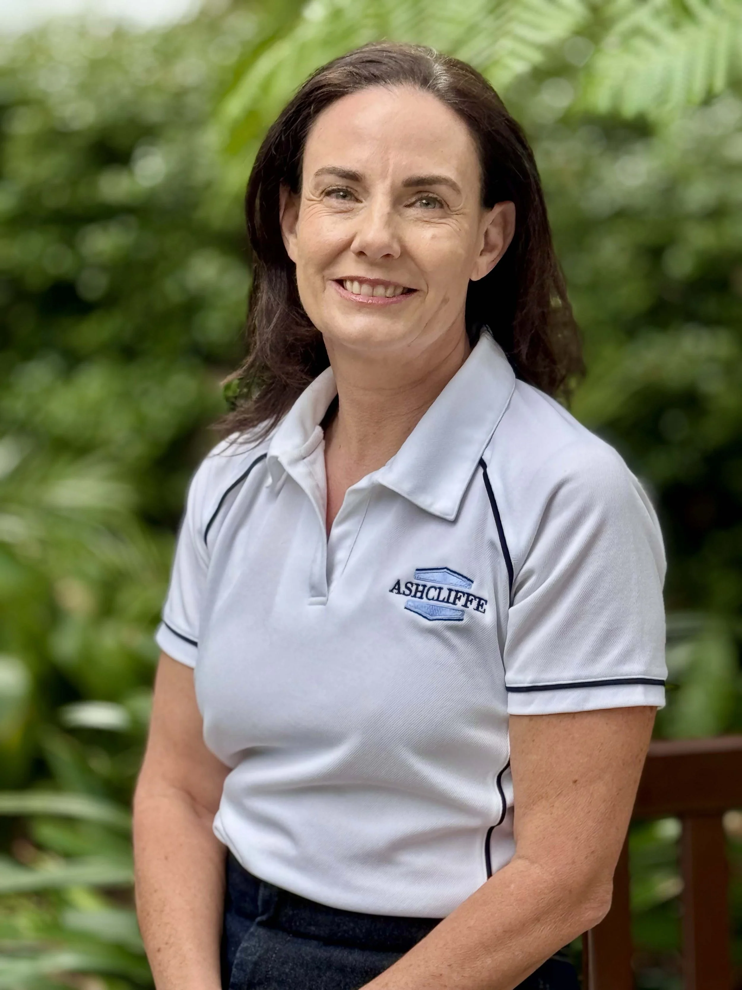 A woman with dark brown hair wearing a white polo shirt with the 'Ashcliff' logo, smiling outdoors with green foliage in the background.