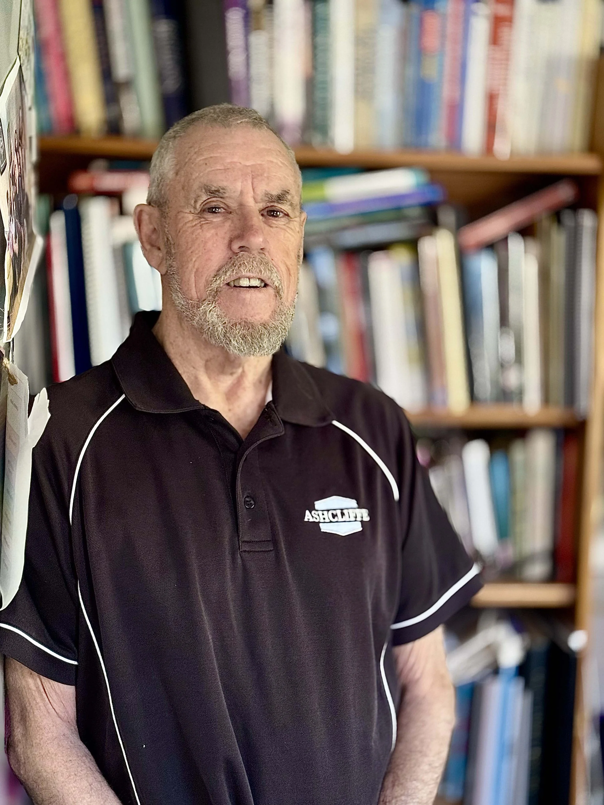 An elderly man with a beard and short hair standing in front of a bookshelf filled with books, wearing a black polo shirt with white piping and the logo 'Ashcliff'.