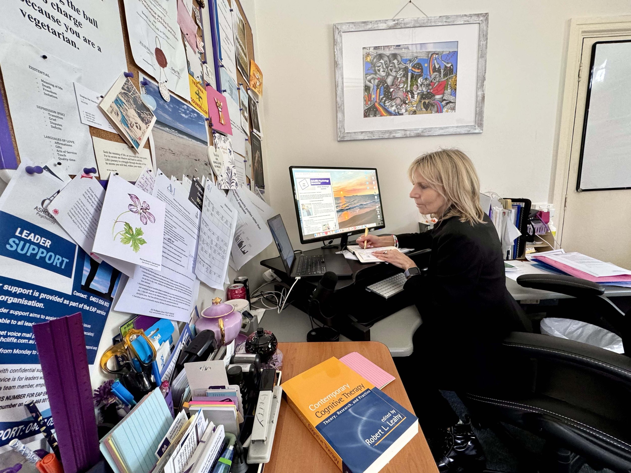 An office with a woman working at a desk, surrounded by papers, notebooks, and office supplies, with a large bulletin board full of papers, notes, and photos on the wall behind her.