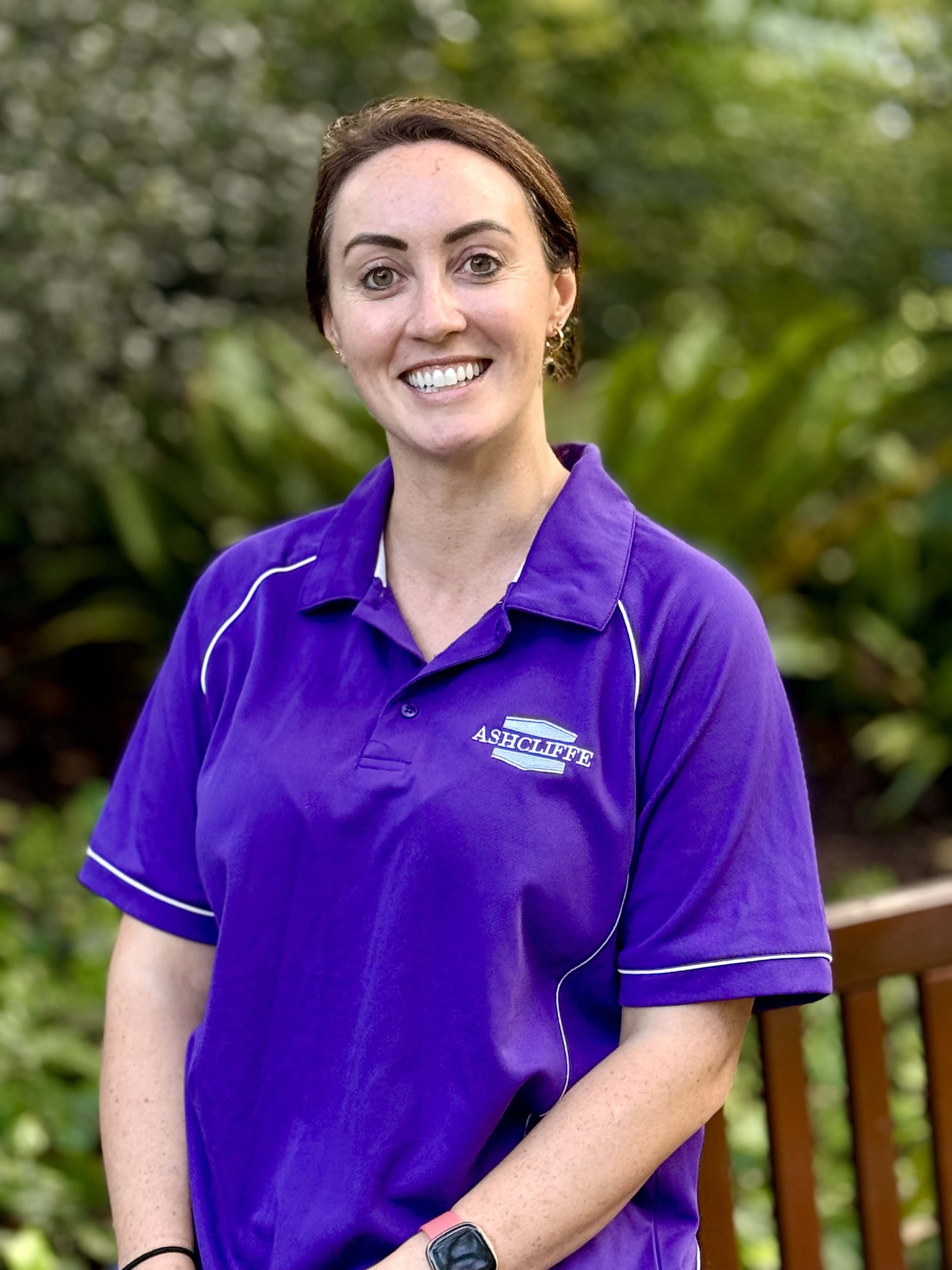 A women with short brunette hair, smiling, wearing a purple sports shirt with 'ASHCLIFFE' embroidered on it, standing outside with greenery in the background.