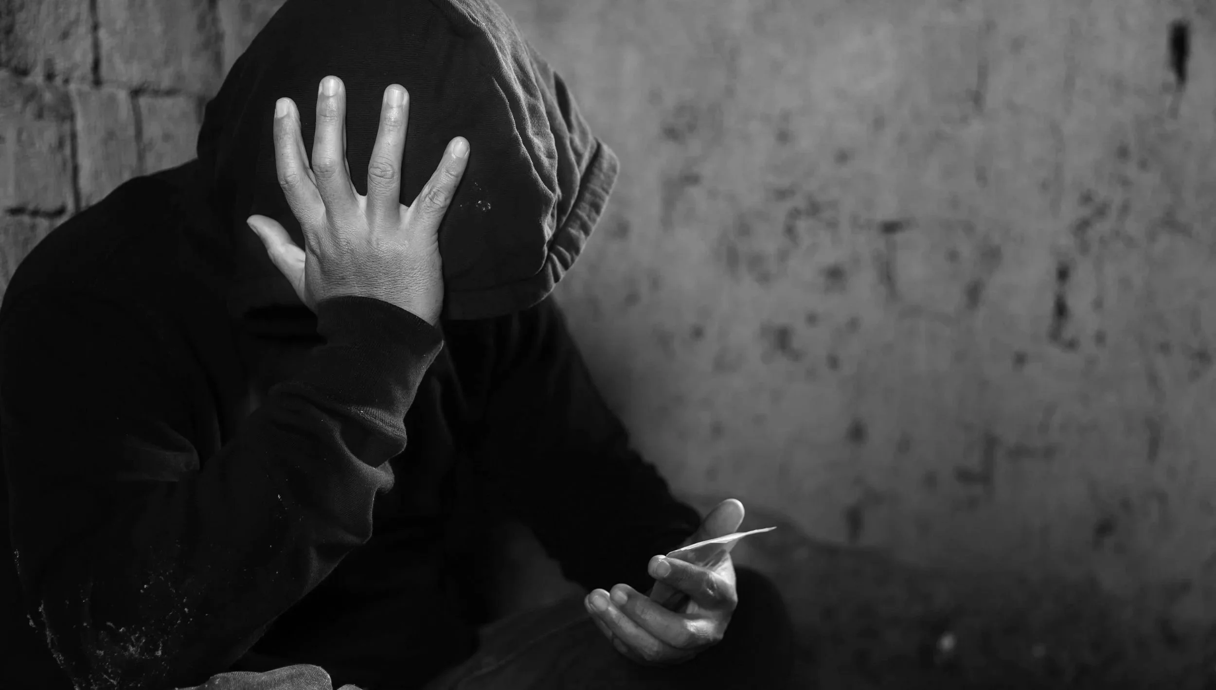 A person wearing a hoodie, sitting against a brick wall, looking at a smartphone, with one hand covering their face.