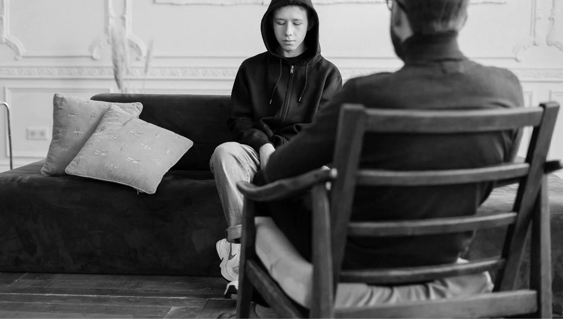 A teenager in a black hoodie sitting on a sofa during an interview or counseling session with an older man in a chair, in a room with ornate wall details.