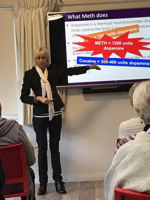woman giving a presentation about methamphetamine effects on the brain with a colorful slide showing dopamine units for meth and cocaine, audience seated in red chairs.