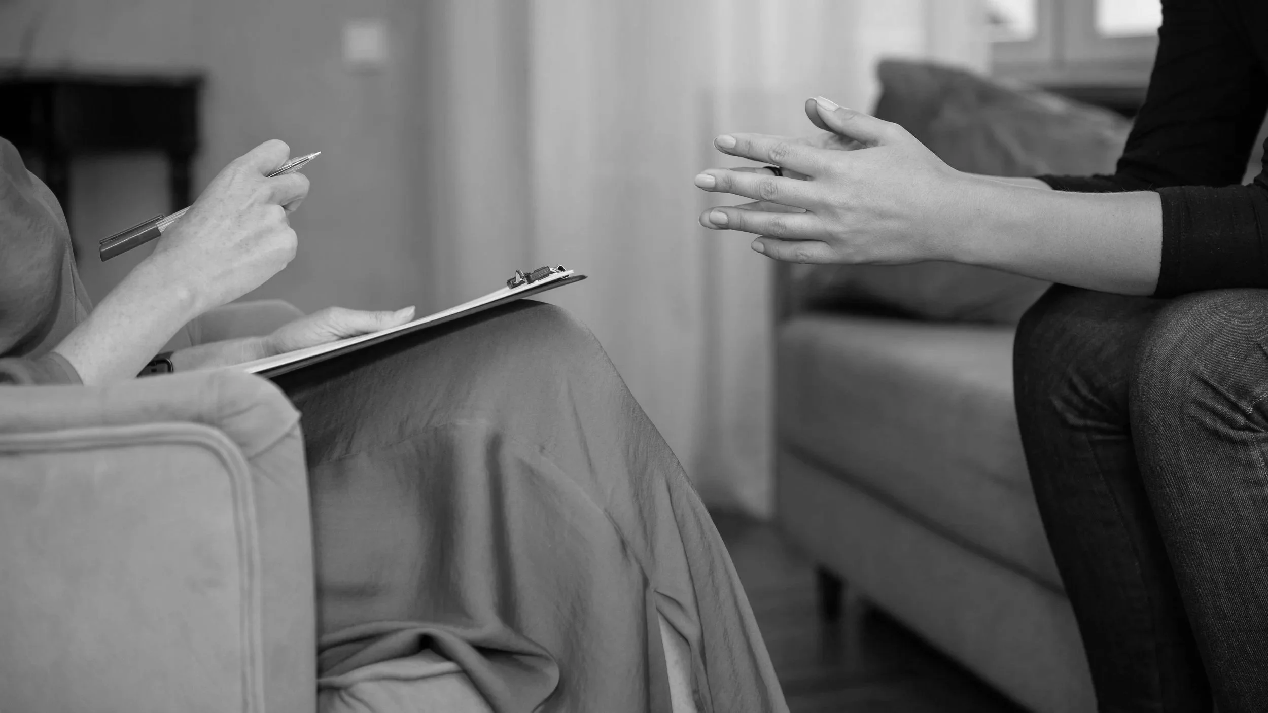 A person sitting on a sofa with a clipboard and pen, talking with another person who is gesturing with their hands during a discussion.