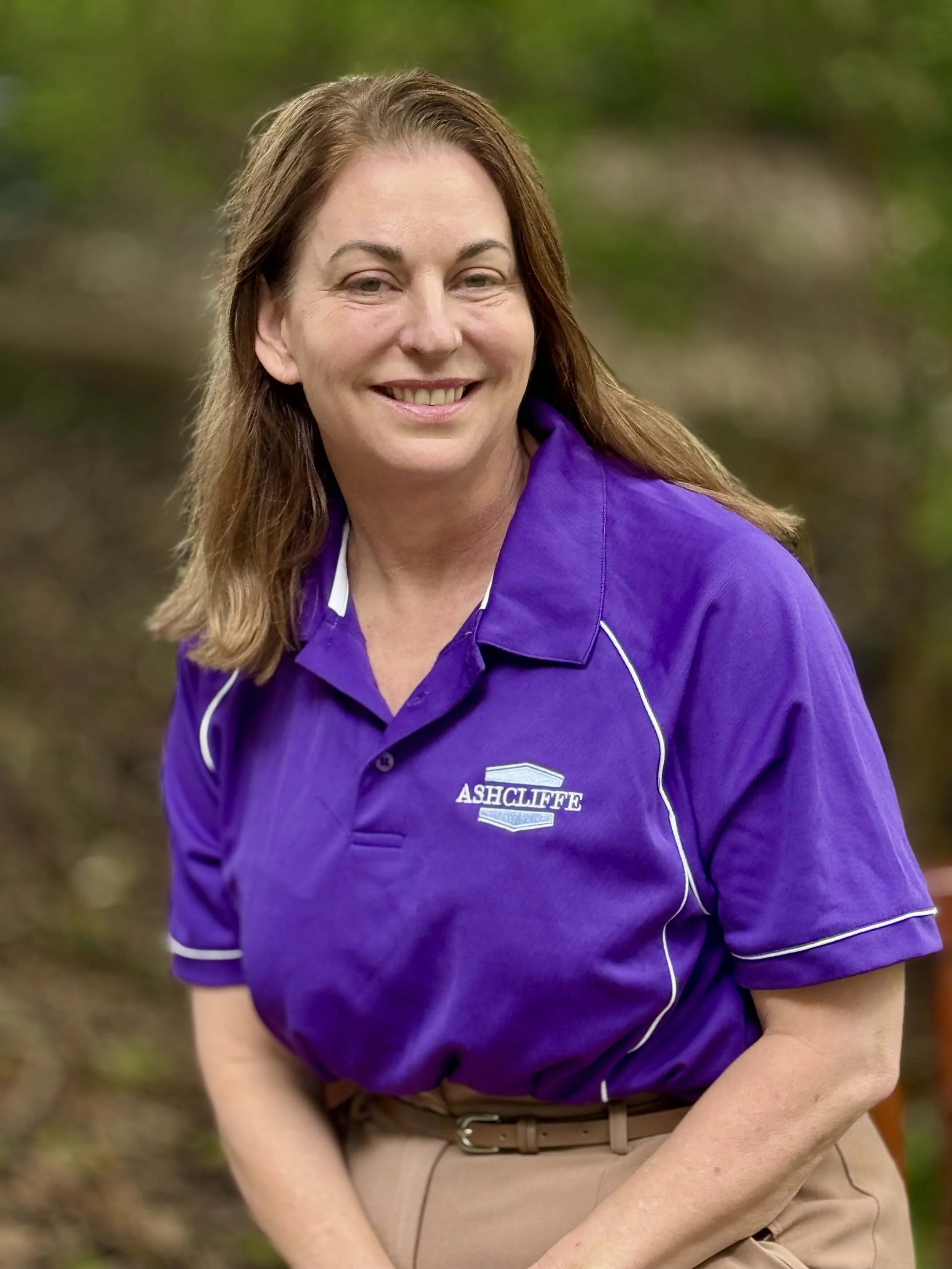 A woman with shoulder-length brown hair smiling, wearing a purple polo shirt with 'ASHCLIFFE' logo, and beige pants, outdoors with blurred greenery in the background.