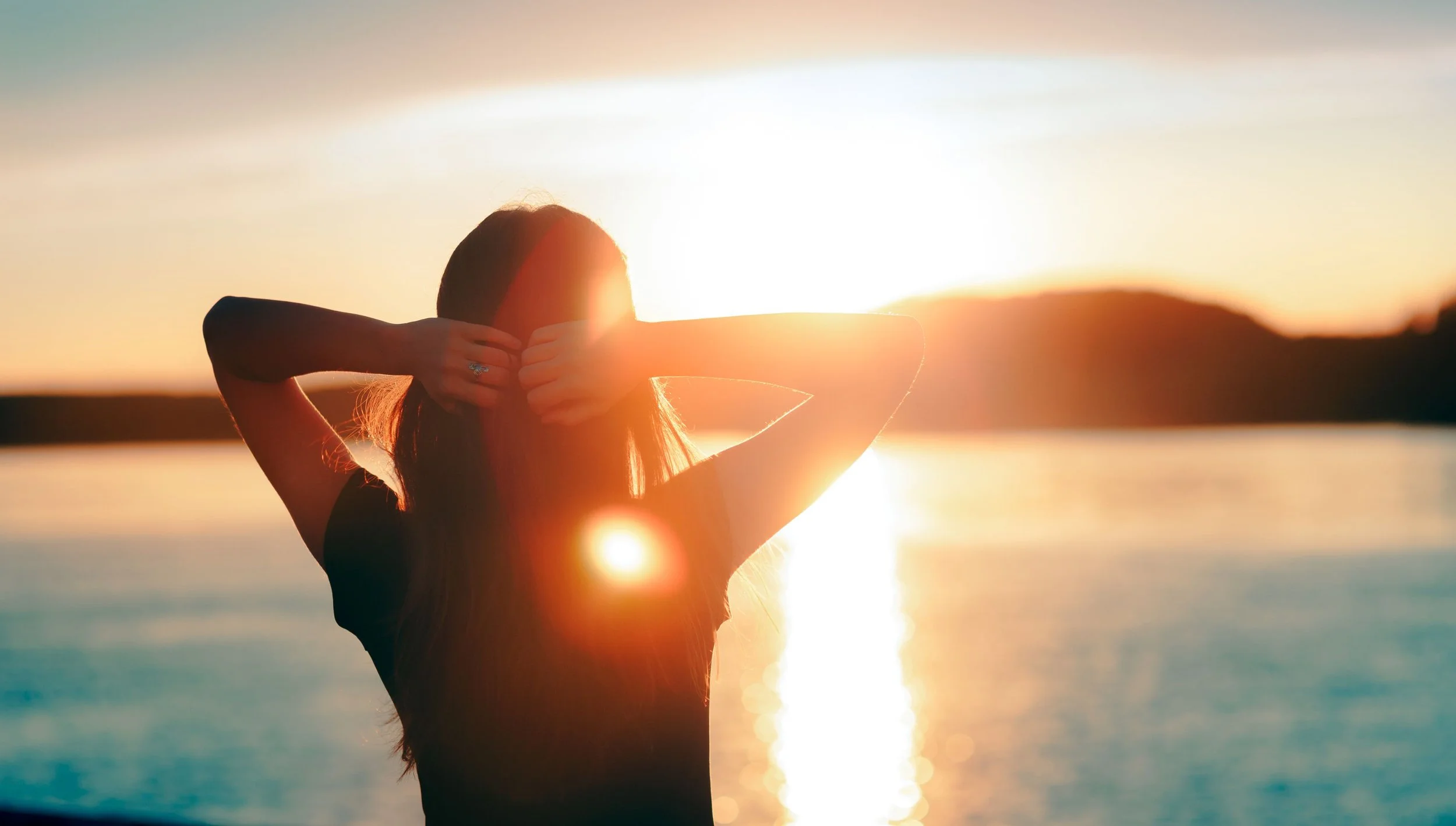 A woman standing outdoors at sunset, with her back to the camera, holding her hair with both hands, near a body of water.