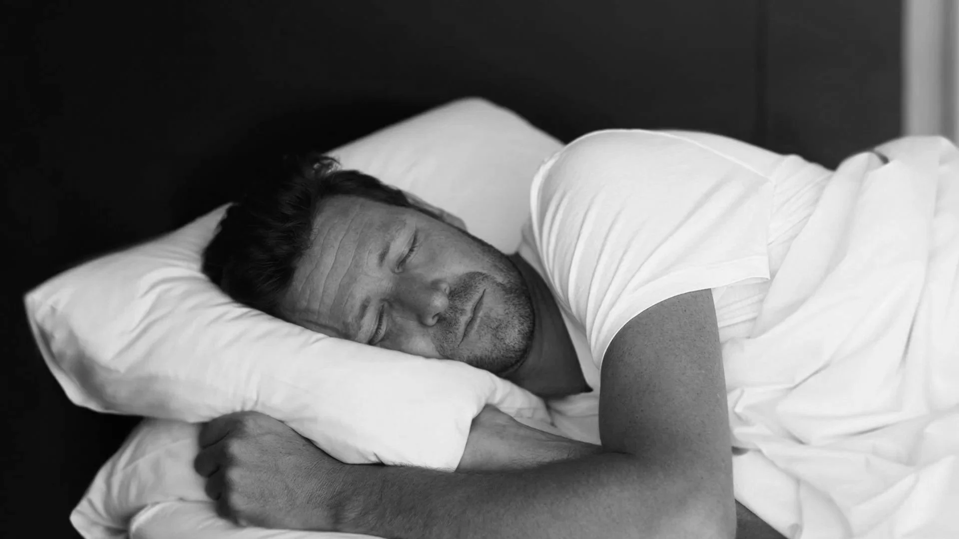 A man sleeping peacefully on his side in bed with a white pillow and white sheets, wearing a white t-shirt.