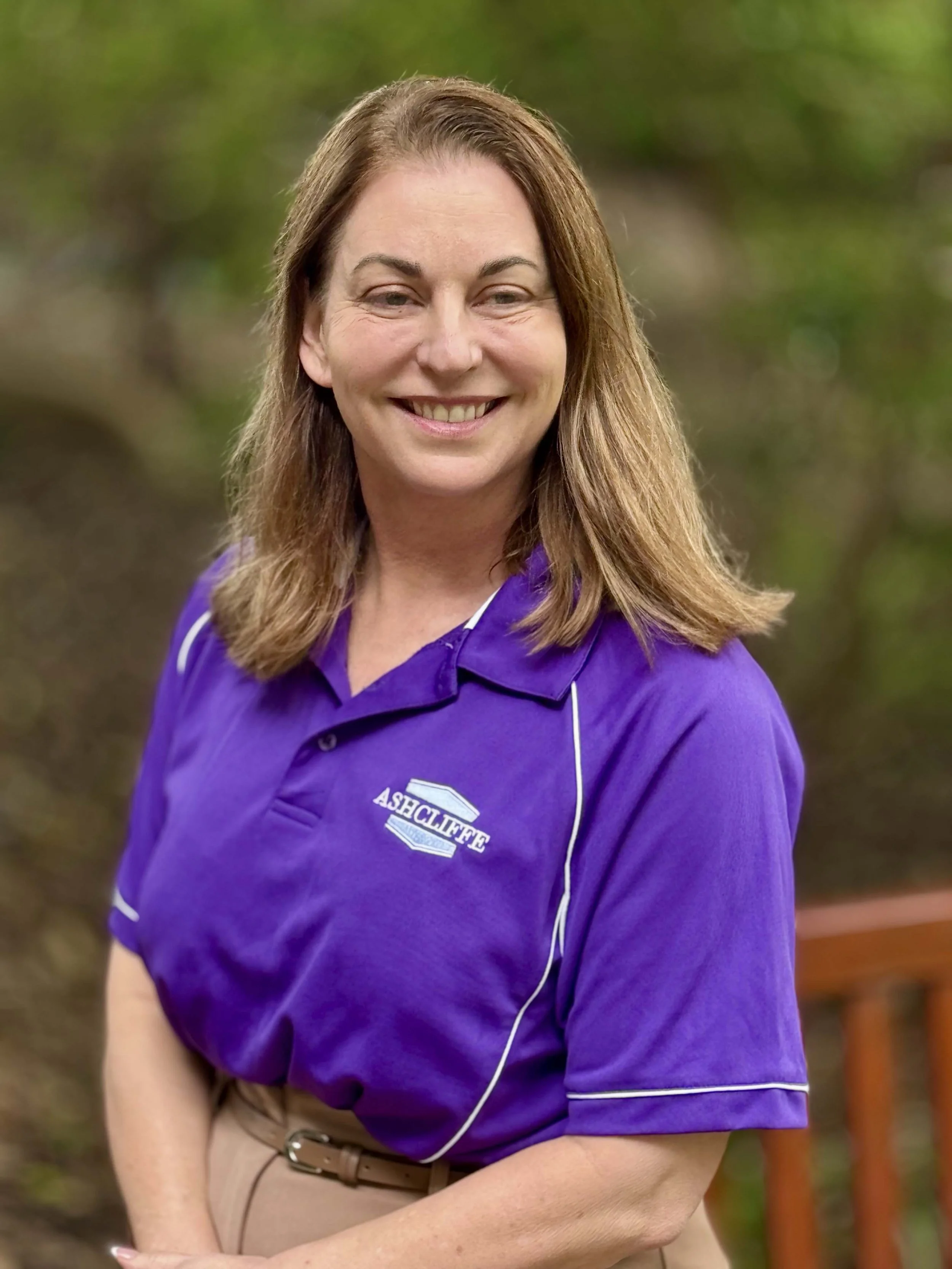 A woman with shoulder-length brown hair, smiling, wearing a purple polo shirt with 'ASHCLIFFE' logo, standing outdoors with greenery in the background.