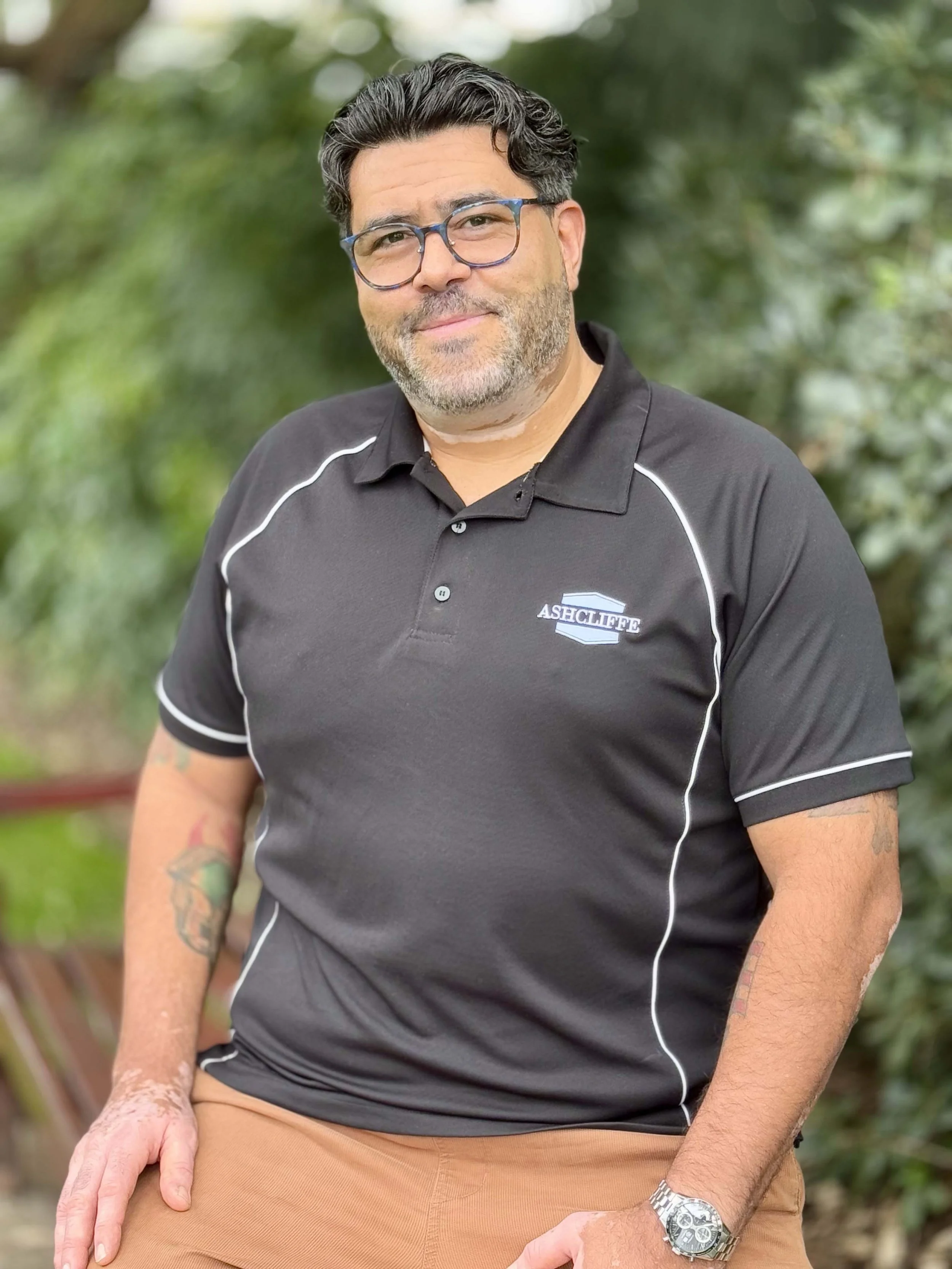 A man with dark hair, glasses, and a beard, wearing a black polo shirt with white trim and the logo 'ASHCLIFFE' on it, sitting outdoors.