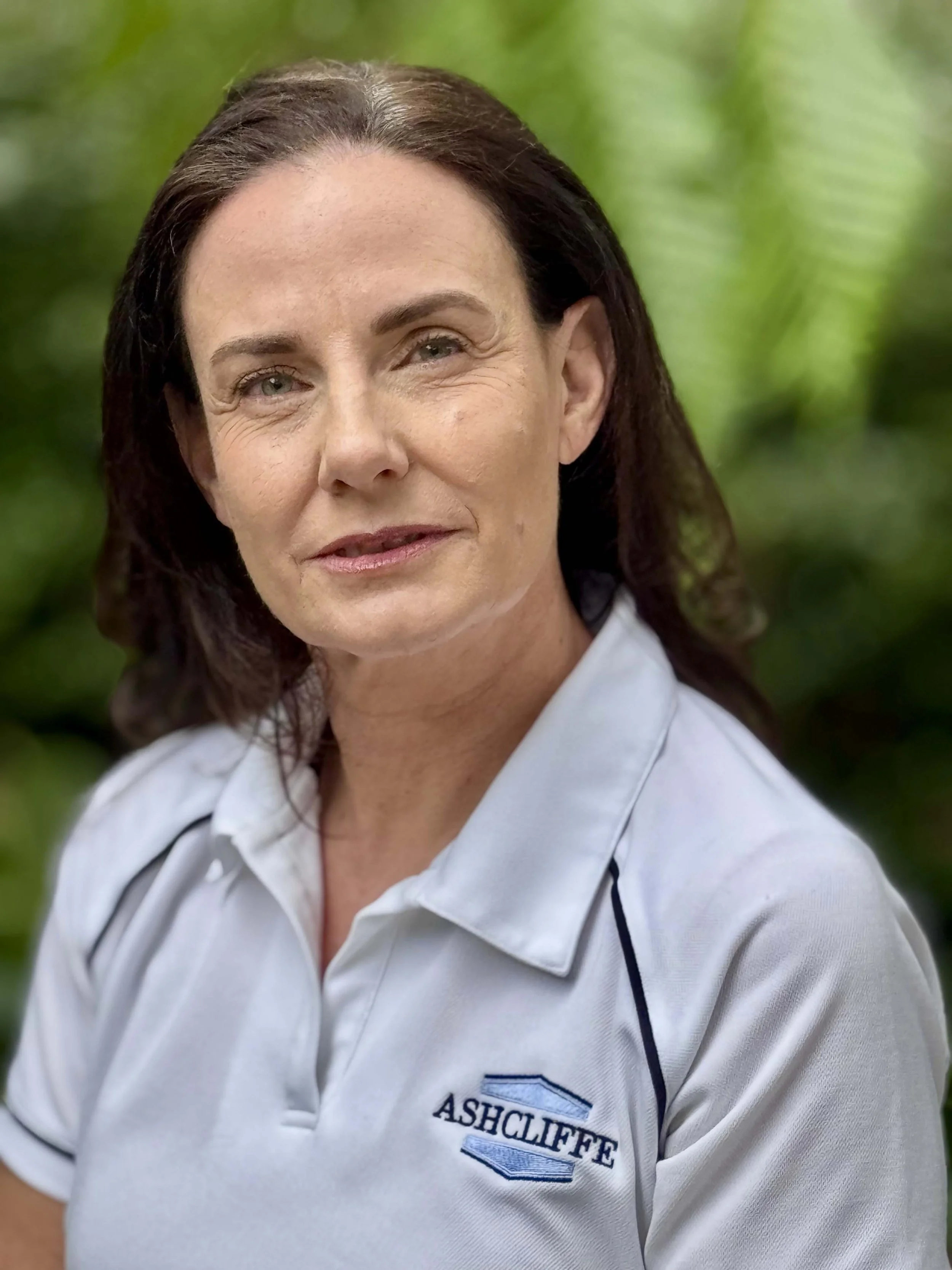 A woman with dark hair wearing a white polo shirt with the 'Ashcliffe' logo on it, standing outdoors with green foliage in the background.