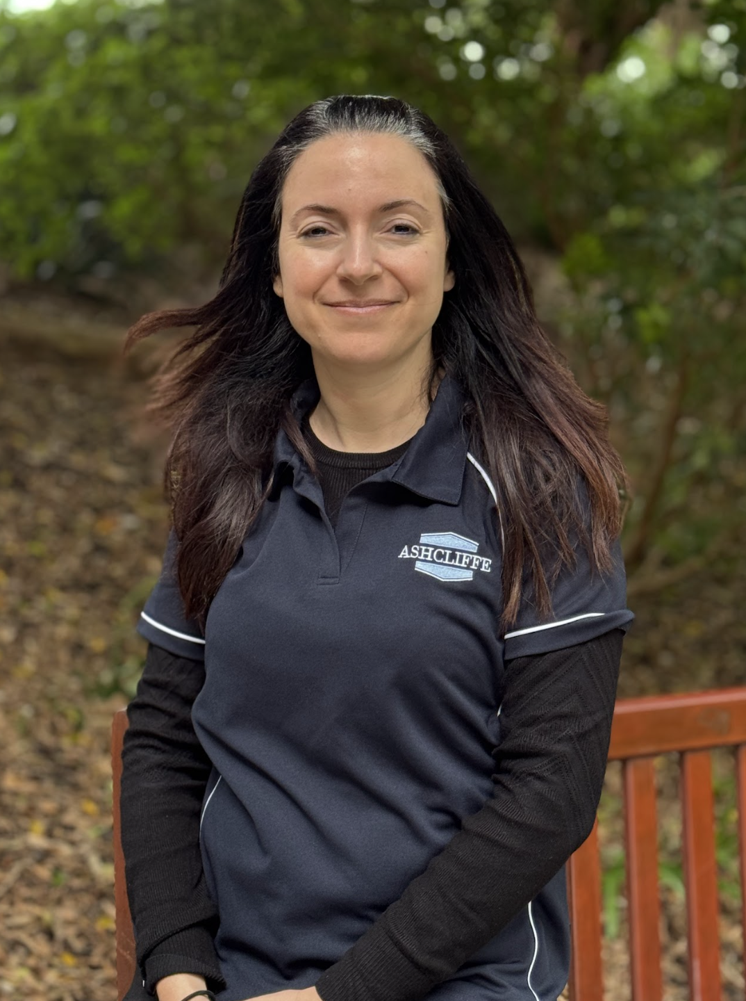 A woman with long dark hair smiling outdoors, wearing a black and blue polo shirt with 'ASHCLIFFE' embroidered on it, in a wooded area with fallen leaves on the ground.