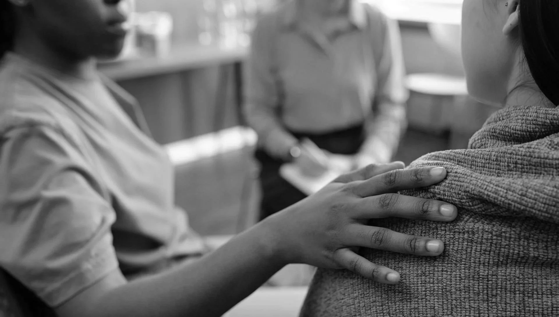 A person with long hair and a textured sweater gently touching the shoulder of another person in a casual setting, with a third person visible in the background.