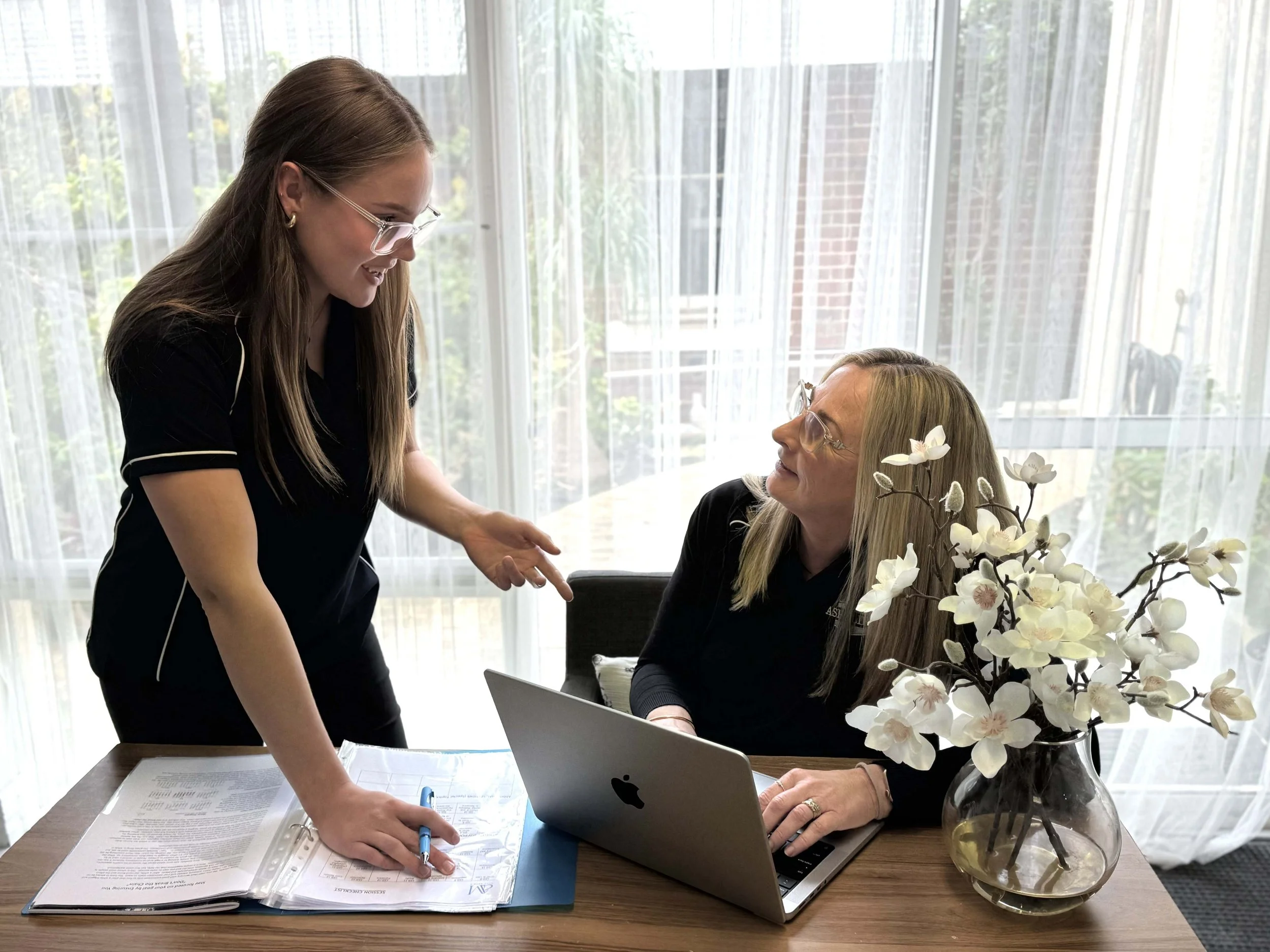 A young woman and an older woman have a friendly discussion at a table, with an open binder and a laptop, in front of large windows with white curtains and a vase of white flowers.