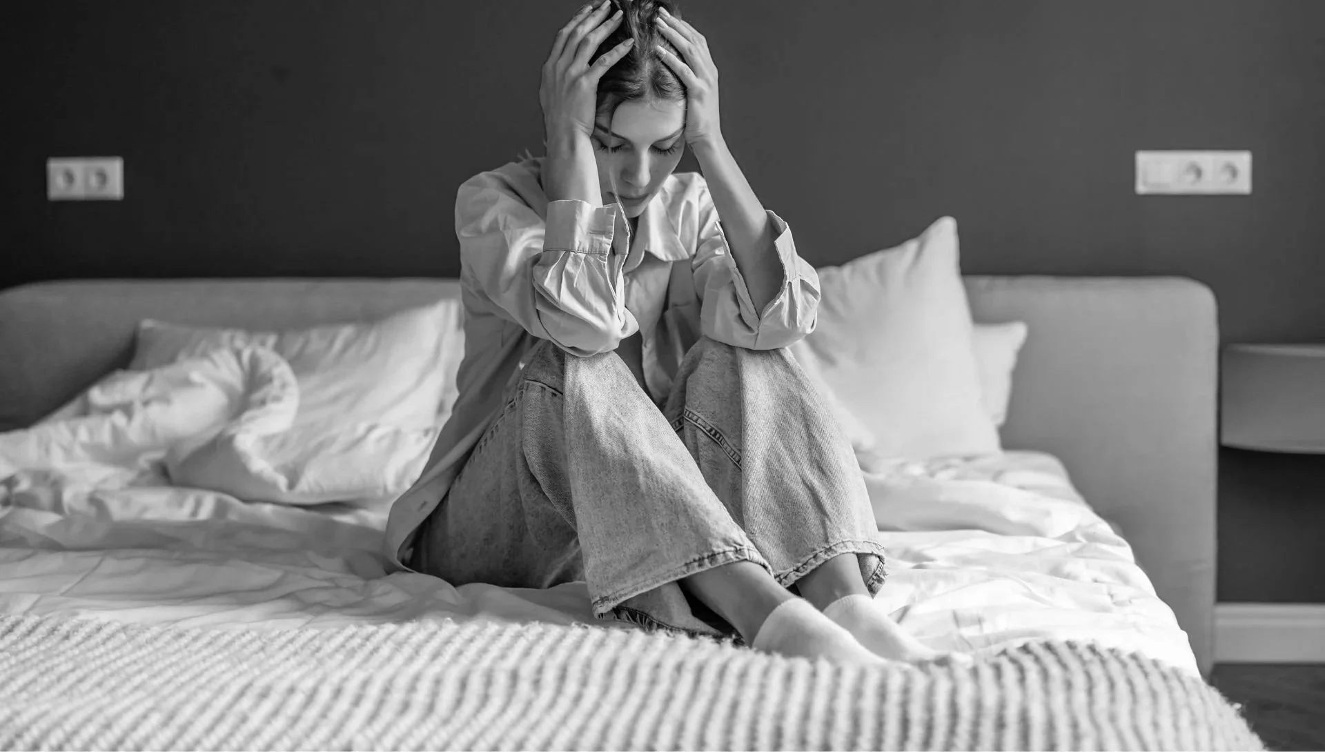 A woman sitting on a bed with her knees drawn up, holding her head in her hands, appearing distressed or upset in a black and white photo.