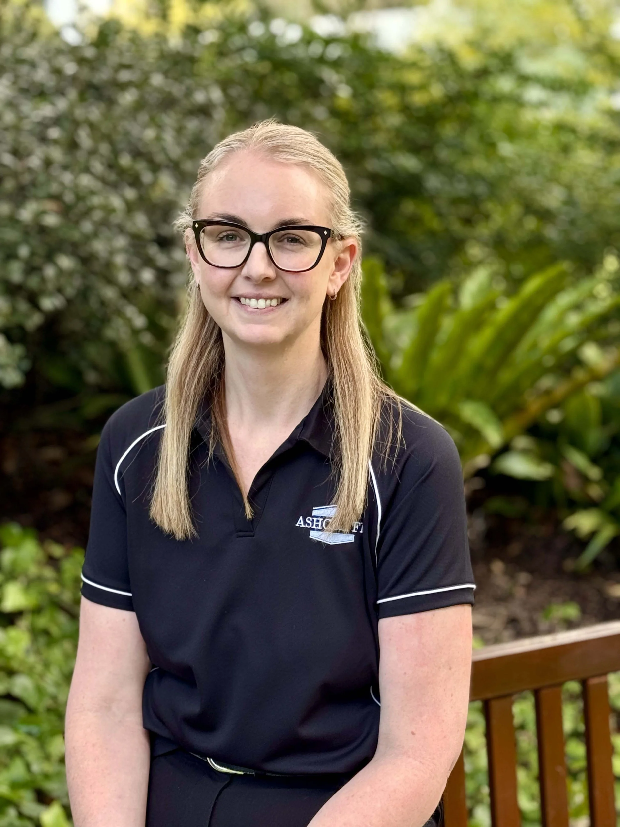 A smiling woman with long blonde hair, wearing glasses and a black polo shirt with a logo, sitting on a wooden bench outdoors with green foliage in the background.
