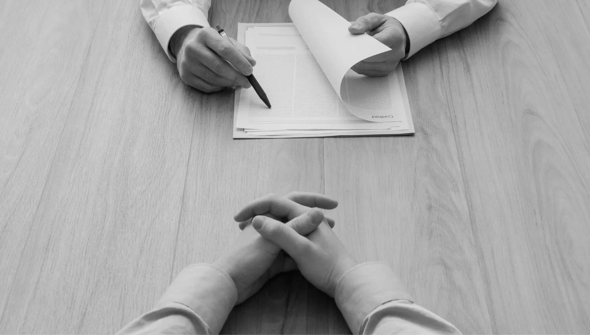 Two people sitting across from each other at a table; one has clasped hands and the other is writing on a document.