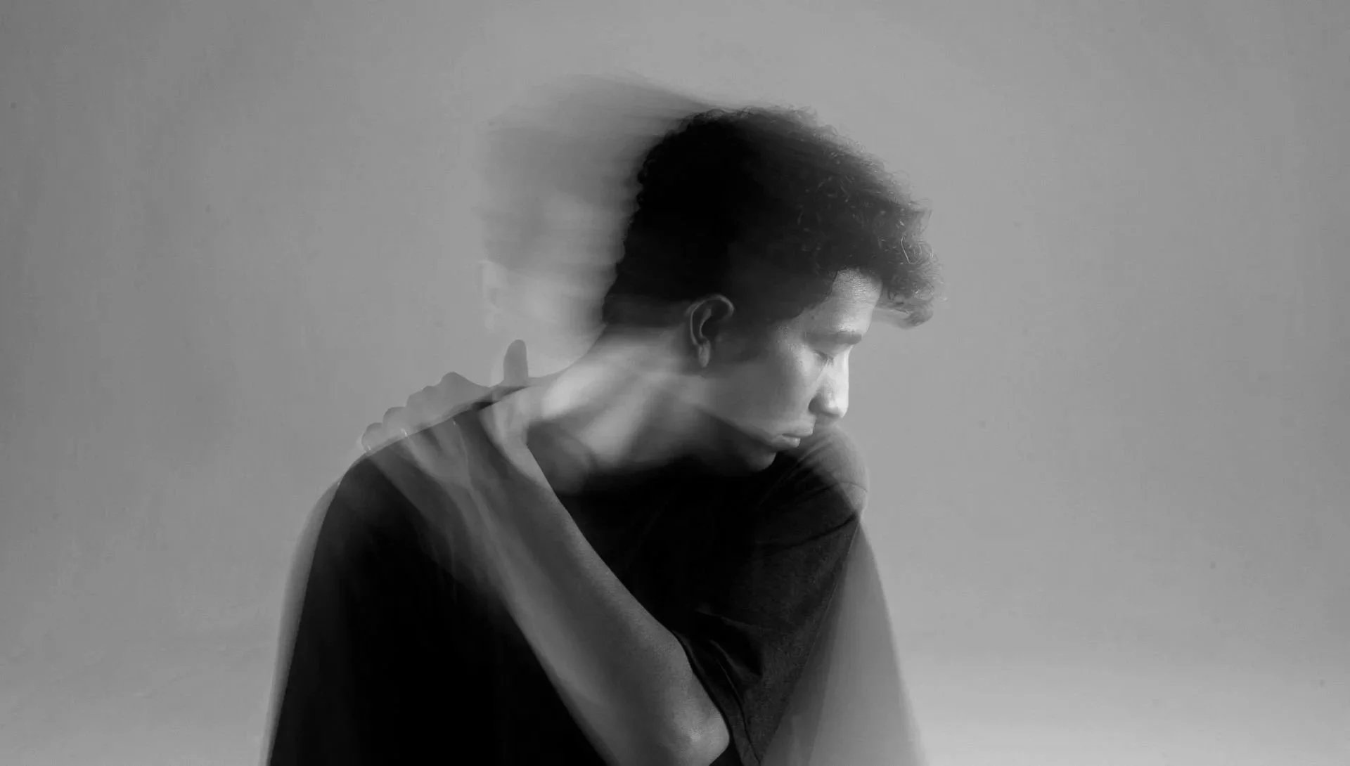 Black and white photo of a person with short curly hair, sitting with their head down and eyes closed, displaying a motion blur effect that creates a ghostly double image.