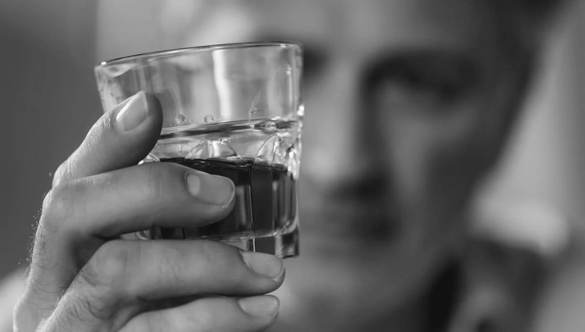 A person holding a glass of water with a finger on the side, out of focus in the background.