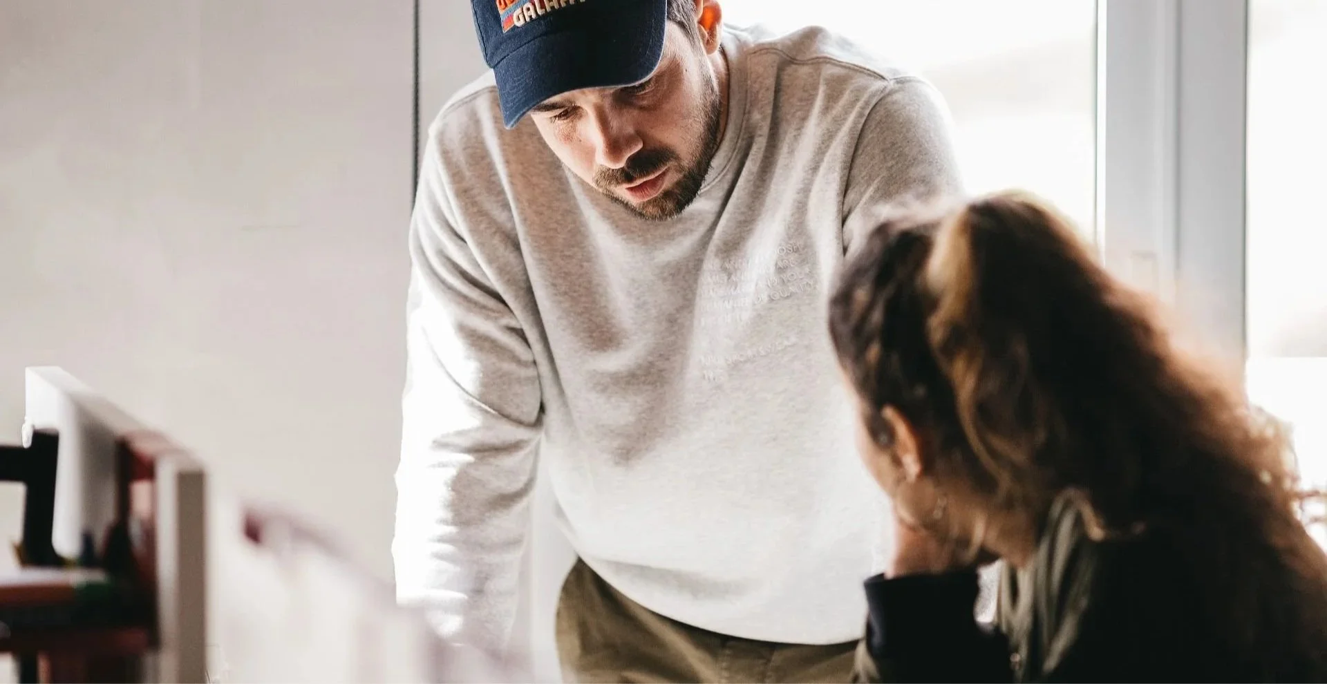 A man and woman having a conversation in a bright indoor setting, with the man leaning over a table and the woman seated.