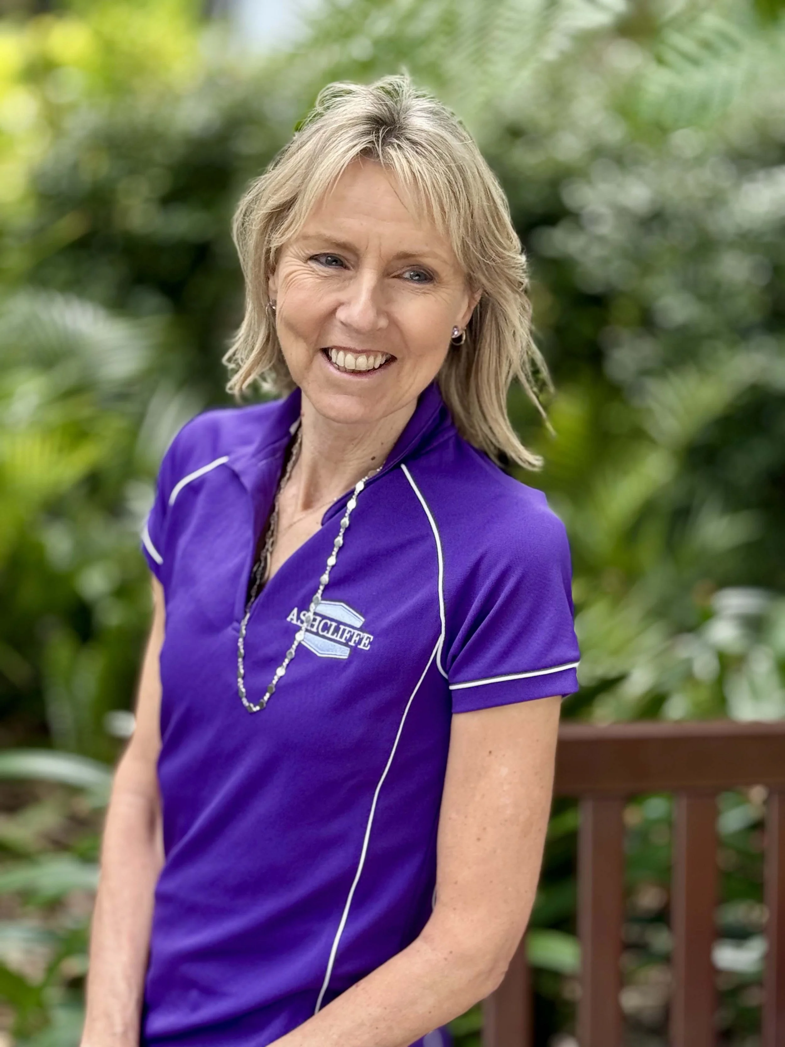 A smiling woman with blonde hair wearing a purple athletic shirt with white piping and a logo that says 'Ashcliff,' standing outdoors with green foliage in the background.
