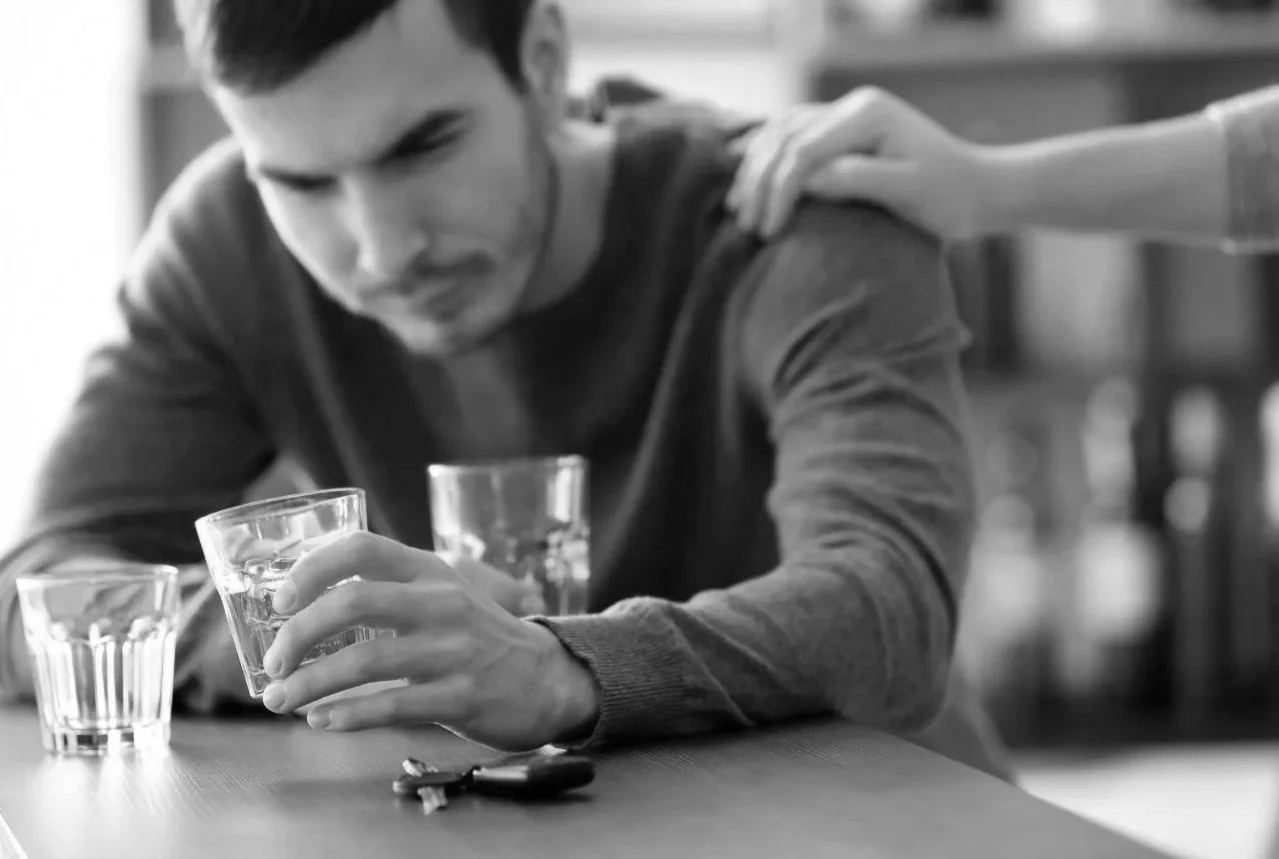 Black and white photo of a man with a worried expression holding a glass of water, with hand on his shoulder, keys on the table, and two empty glasses in front.