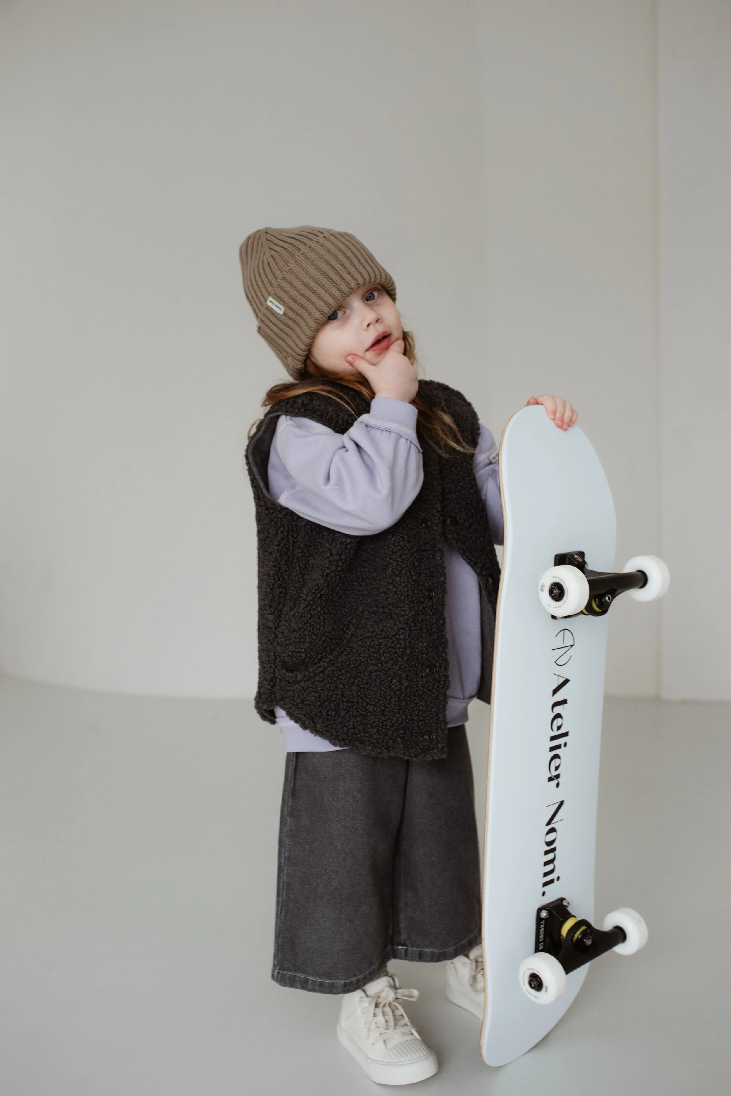 Ein kleines Mädchen mit braunem Strickmütze, grauem Pullover, schwarzem Fleecepullover und grauen Hosen, das aufrecht steht und einen weißen Skateboard hält.