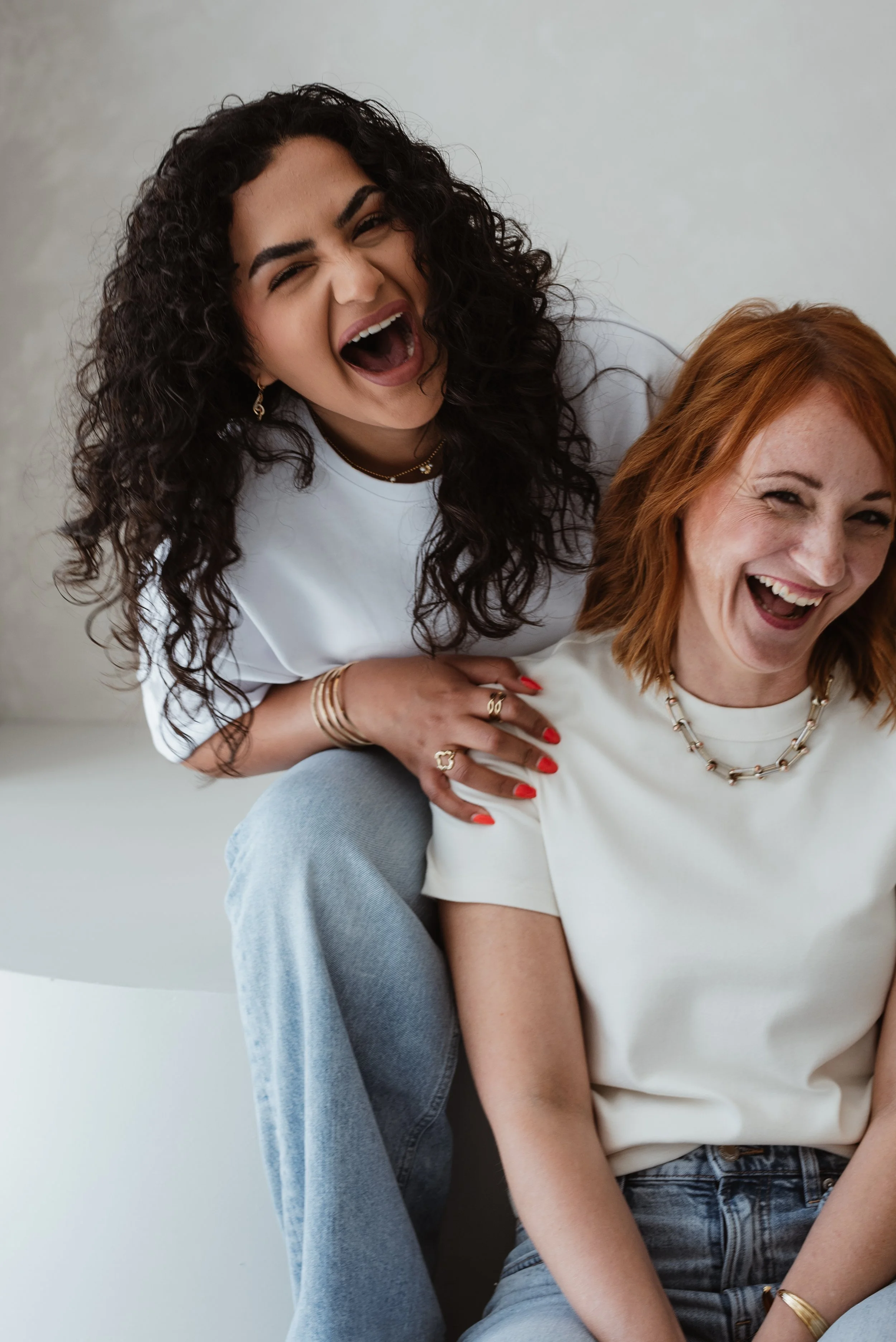 Zwei lachende Frauen sitzen eng beieinander, eine traut sich mit offenem Mund zu lachen, die andere lächelt breit, beide tragen Schmuck und legere Kleidung.