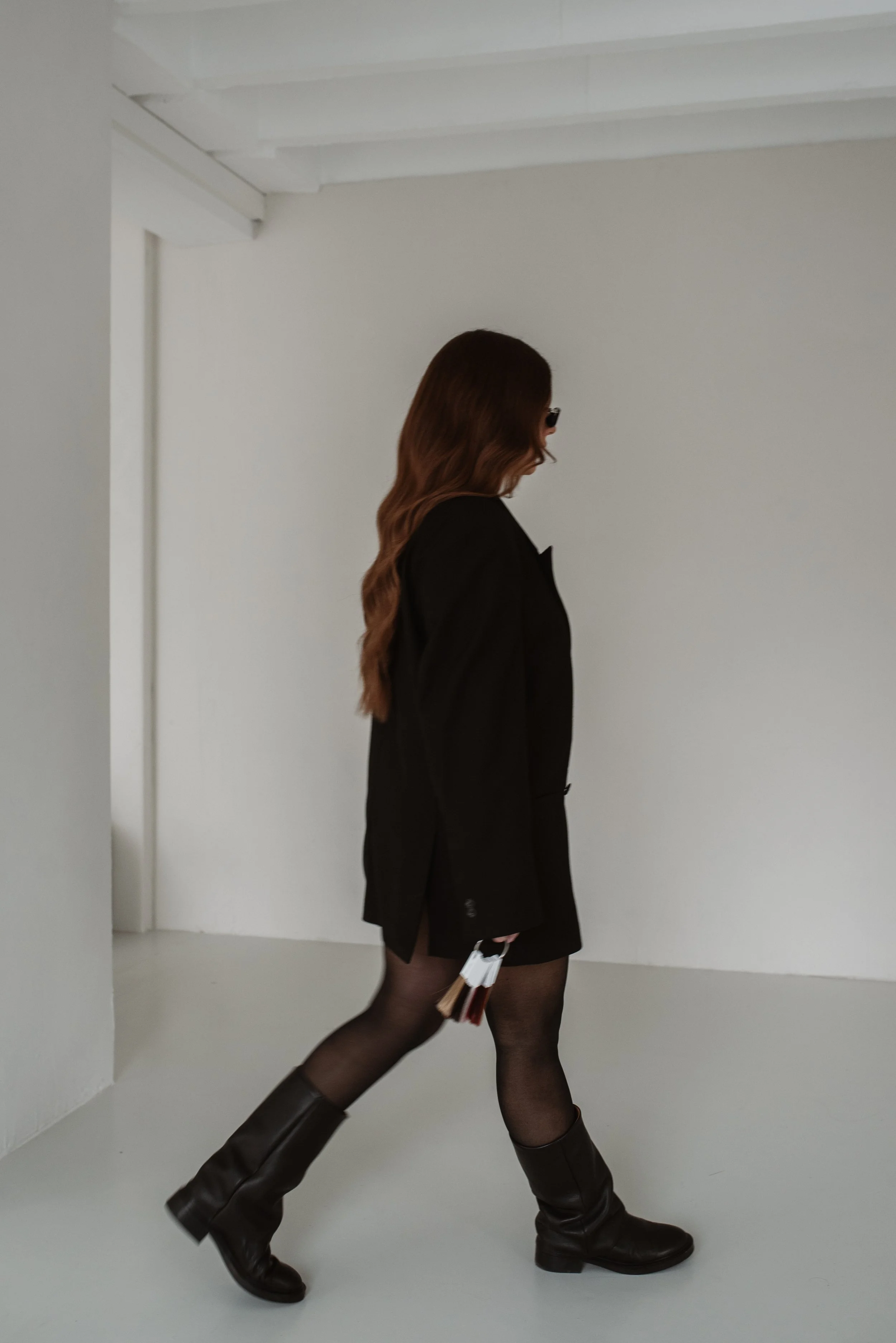 Eine Frau mit langen braunen Haaren trägt einen schwarzen Mantel, einen schwarzen Rock, schwarze Strumpfhosen und braune Stiefel, während sie in einem weißen Raum geht.