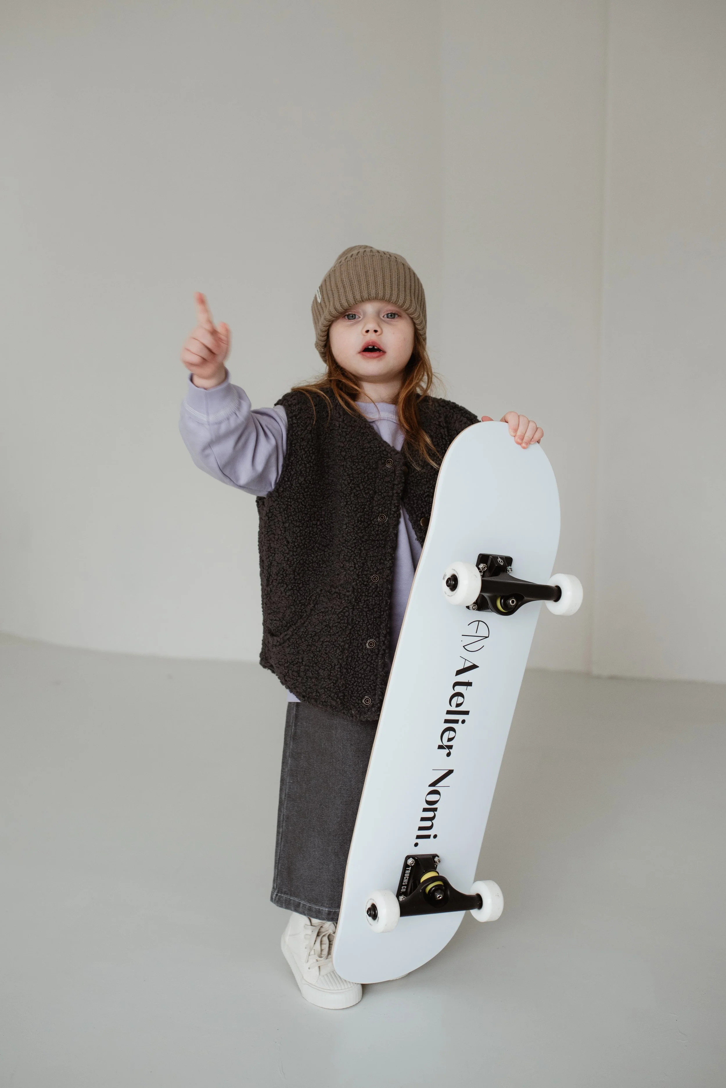 Ein Mädchen trägt eine braune Mütze, eine schwarze Weste, ein lila Oberteil, eine graue Wollhose und Weißschuhe. Es steht auf einem weißen Skateboard mit schwarzen Rädern, hält es mit der linken Hand und zeigt mit dem rechten Zeigefinger nach oben.