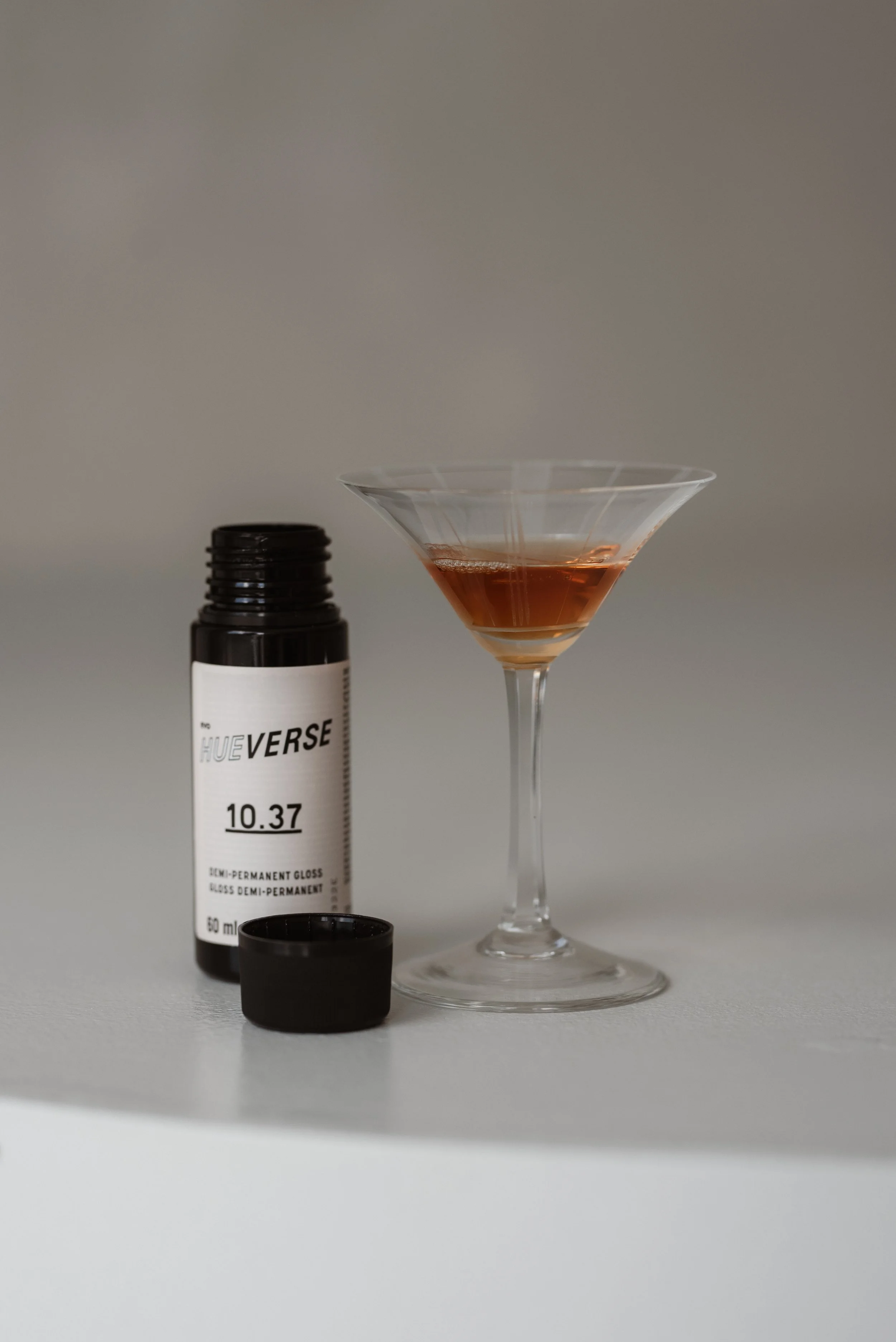 Ein Cocktail in einem Martini-Glas, neben einer schwarzen Flasche mit weißem Etikett und einem schwarzen Verschluss, alle auf einer weißen Oberfläche.