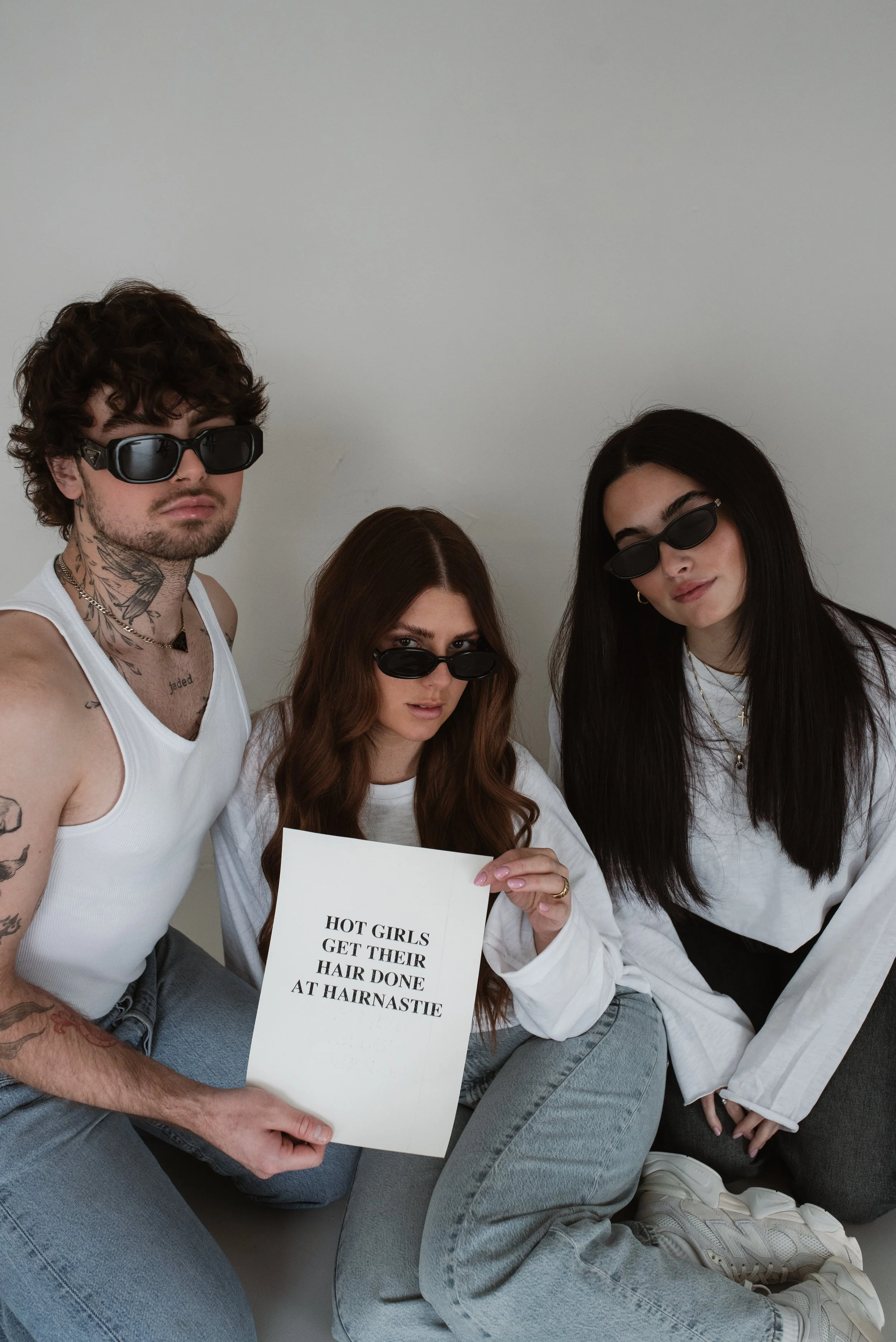 Drei junge Menschen in weißen T-Shirts und Sonnenbrillen sitzen auf dem Boden. Die Person in der Mitte hält ein Schild mit der Aufschrift 'HOT GIRLS GET THEIR HAIR DONE AT HAIRNASTIE'.