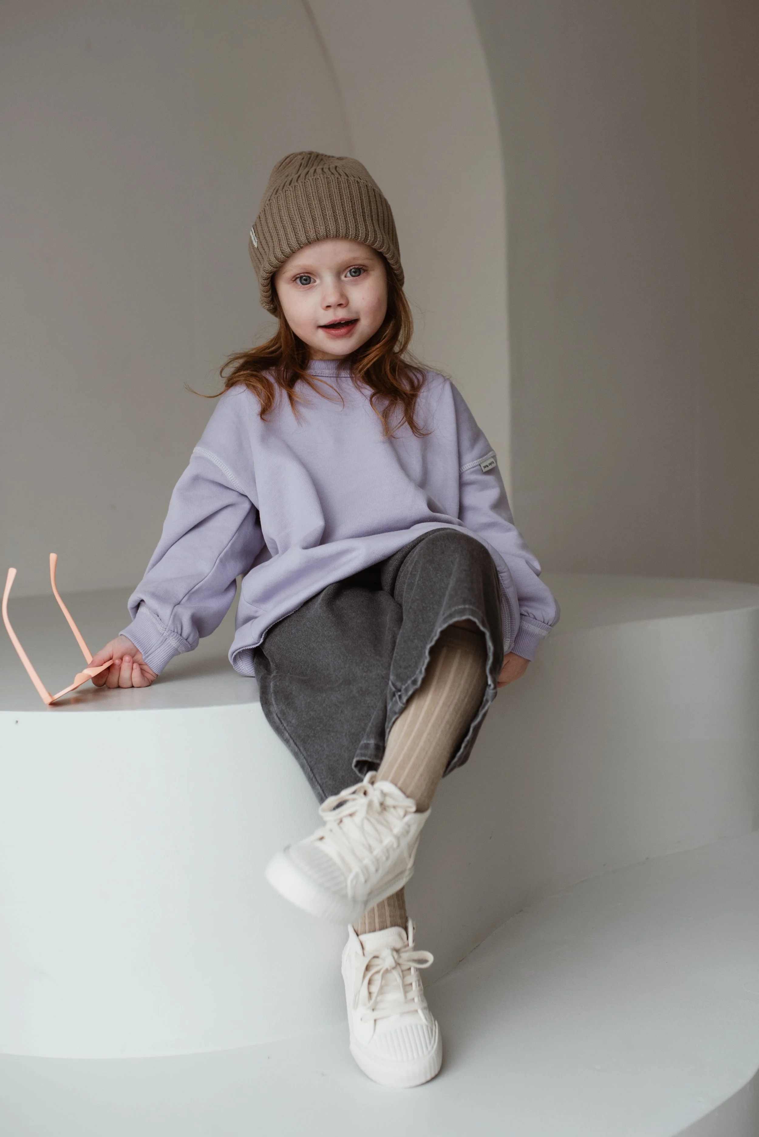 Ein junges Mädchen sitzt auf einer weißen Plattform, trägt einen braunen Wollmützen, ein lila Hemd, graue Hosen, beige Strumpfhosen und weiße Turnschuhe. Sie hält ein rosa Objekt in der Hand und schaut in die Kamera.
