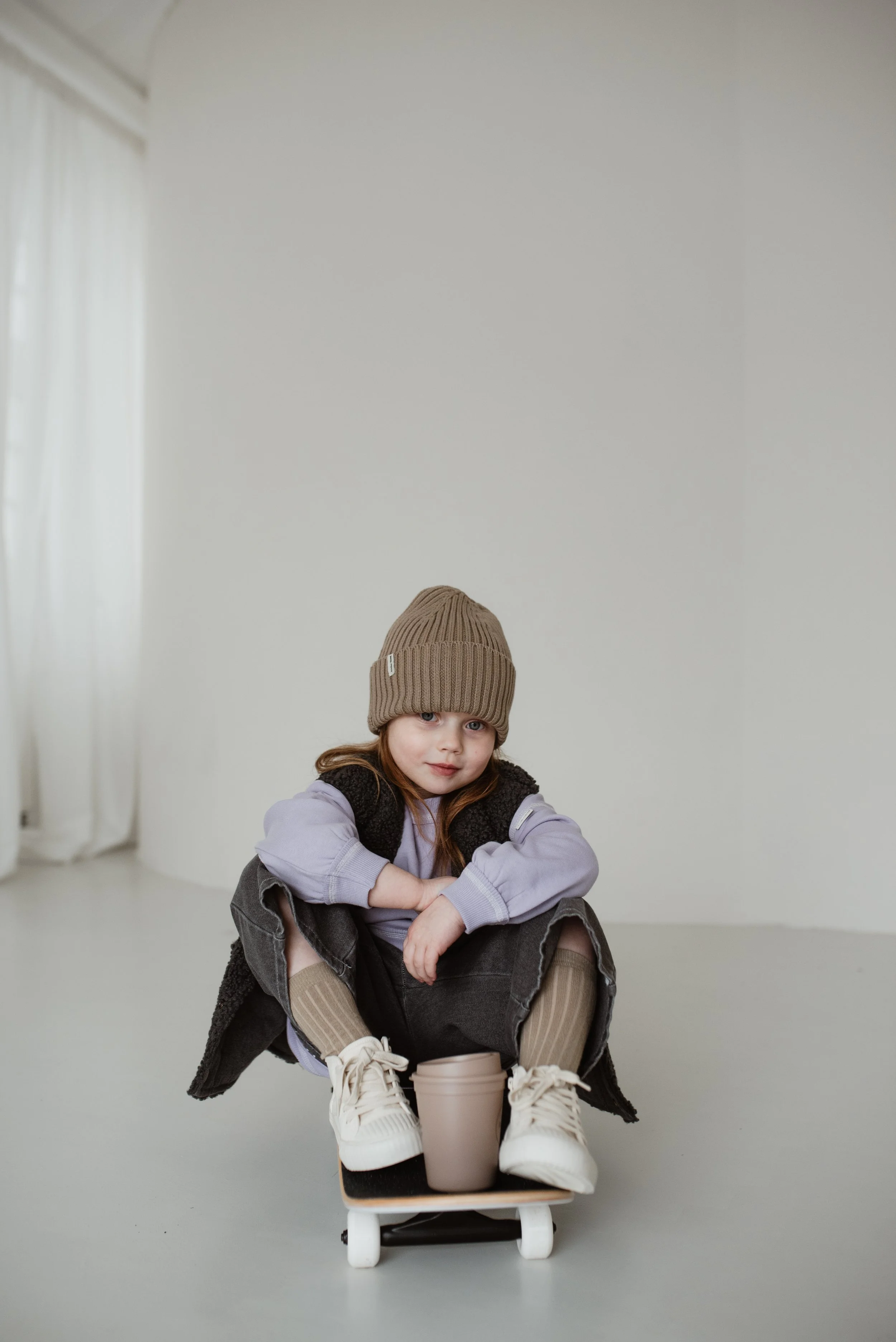 Ein Mädchen in Mütze und Mantel sitzt auf einem Skateboard, verschränkt die Arme und schaut in die Kamera. Im Vordergrund befindet sich eine Tasse.