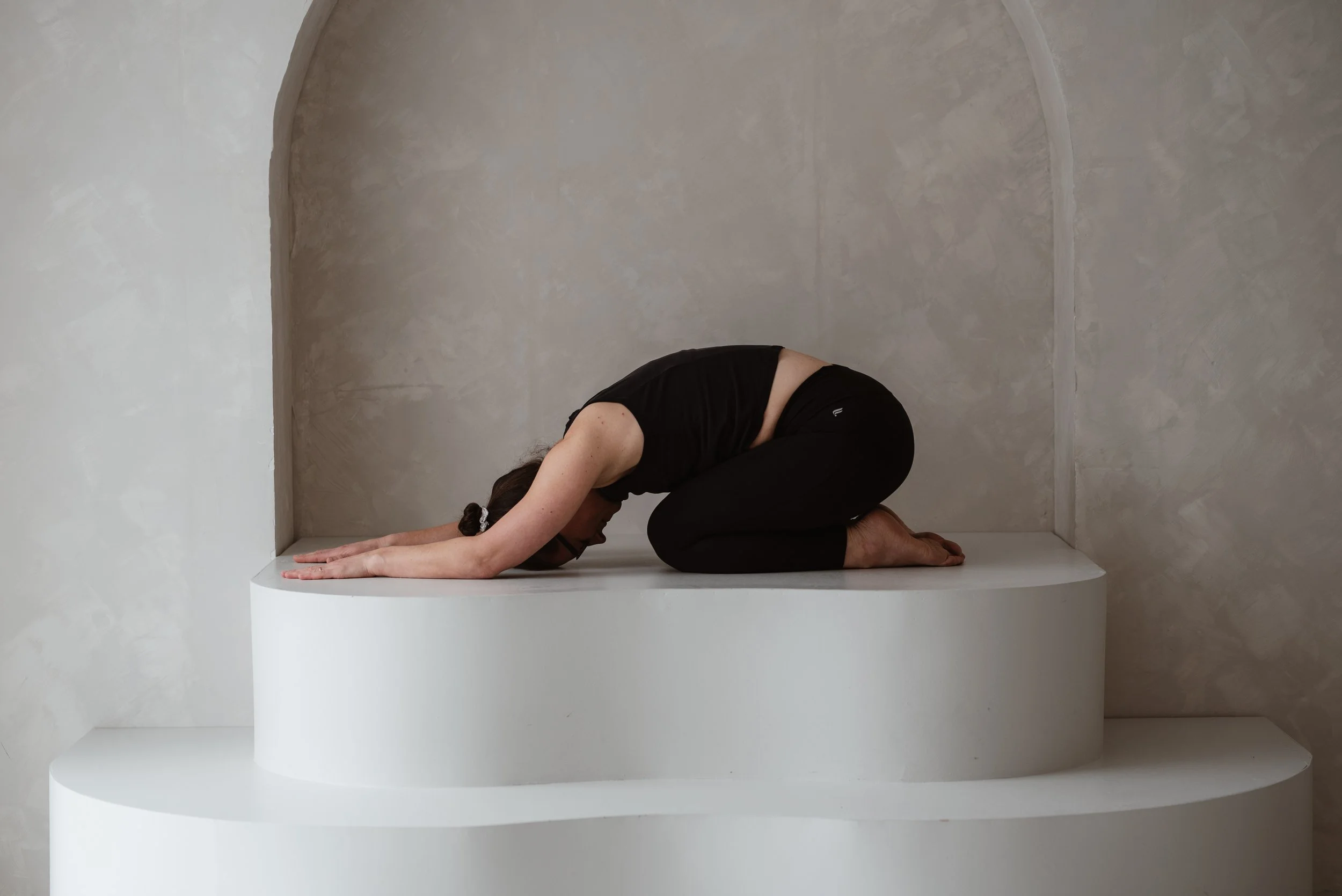 Frau macht eine Yoga-Position, kniet und beugt sich mit den Händen flach auf den Boden, in einem minimalistischen Raum mit weißen Wänden und einer Plattform.
