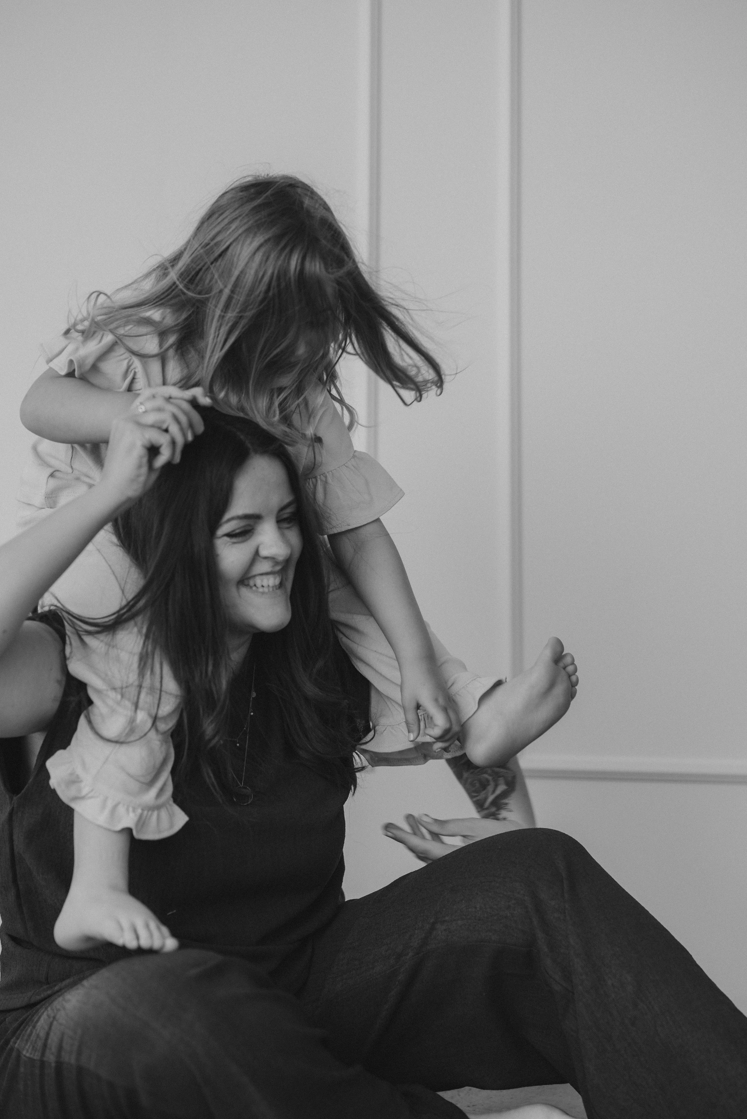 Eine lachende Frau mit dunklen Haaren trägt ein Kind auf den Schultern, das fröhlich lacht. Sie sitzen im Innenraum, wahrscheinlich in einem Raum ohne besonderen Hintergrund.