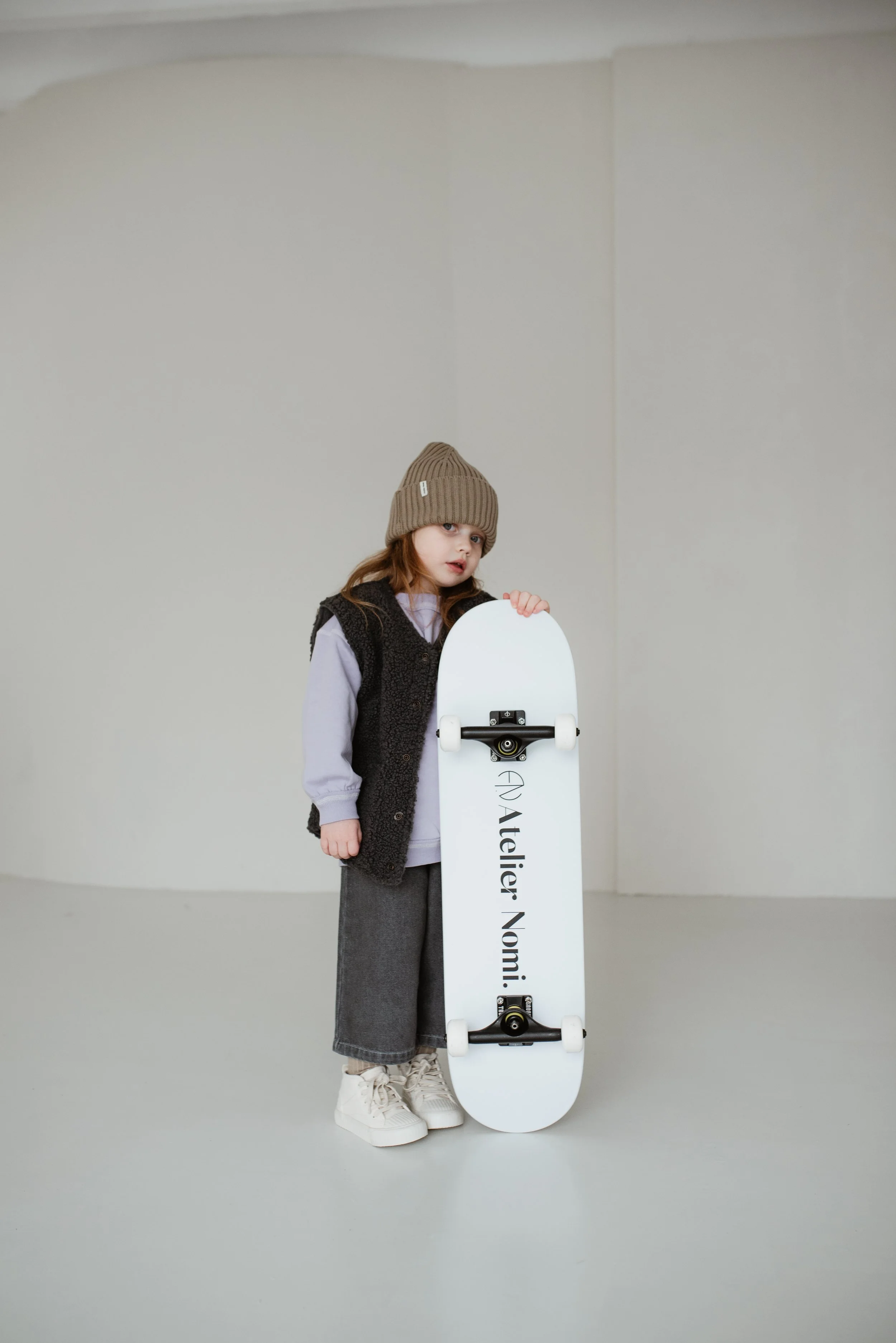 Ein Mädchen mit braunem Wollhut, grauer Hose, weißem T-Shirt und schwarzer Weste steht mit einem weißen Skateboard vor einer hellen, leeren Wand.