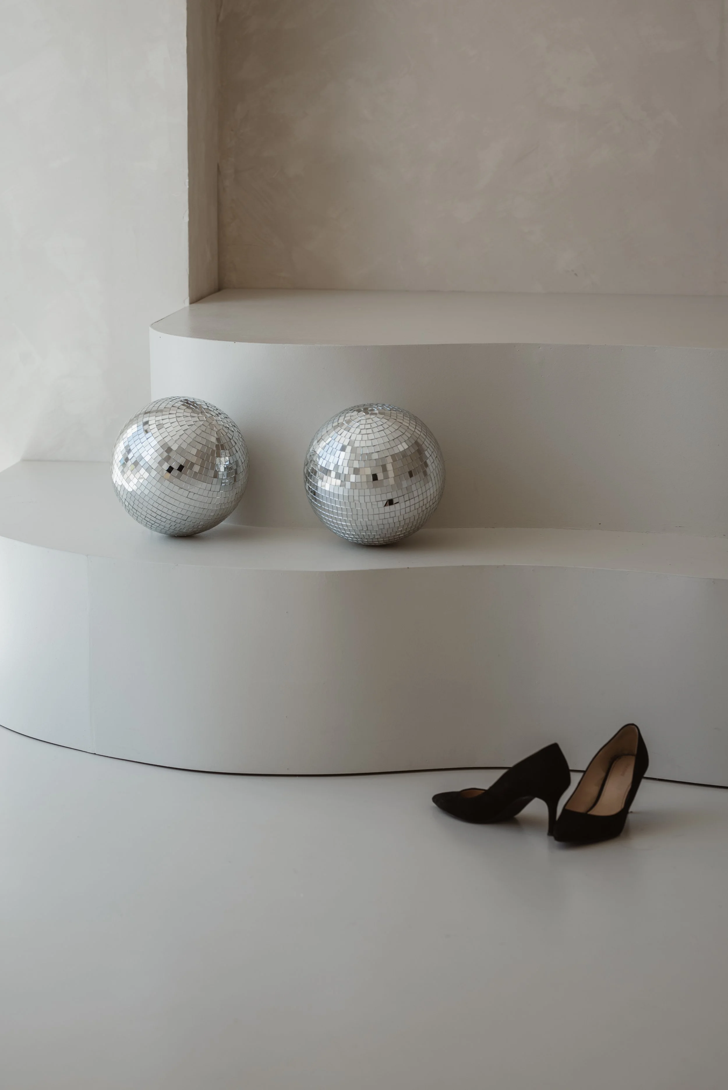 Schwarze High Heels stehen auf dem Boden neben einem weißen Möbelstück. Auf dem Möbelstück sind zwei dekorative Spiegelkugeln.