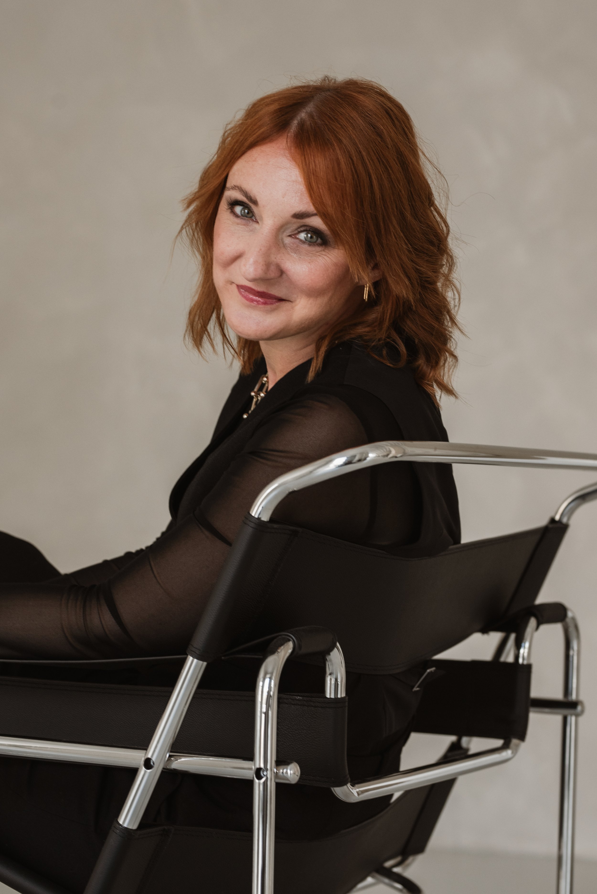 Das Bild zeigt eine Frau mit roten, lockigen Haaren, die auf einem schwarzen Rollstuhl sitzt. Sie trägt einen schwarzen, langärmligen, transparenten Top. Sie lächelt und blickt leicht zur Kamera, vor einem grauen Hintergrund.