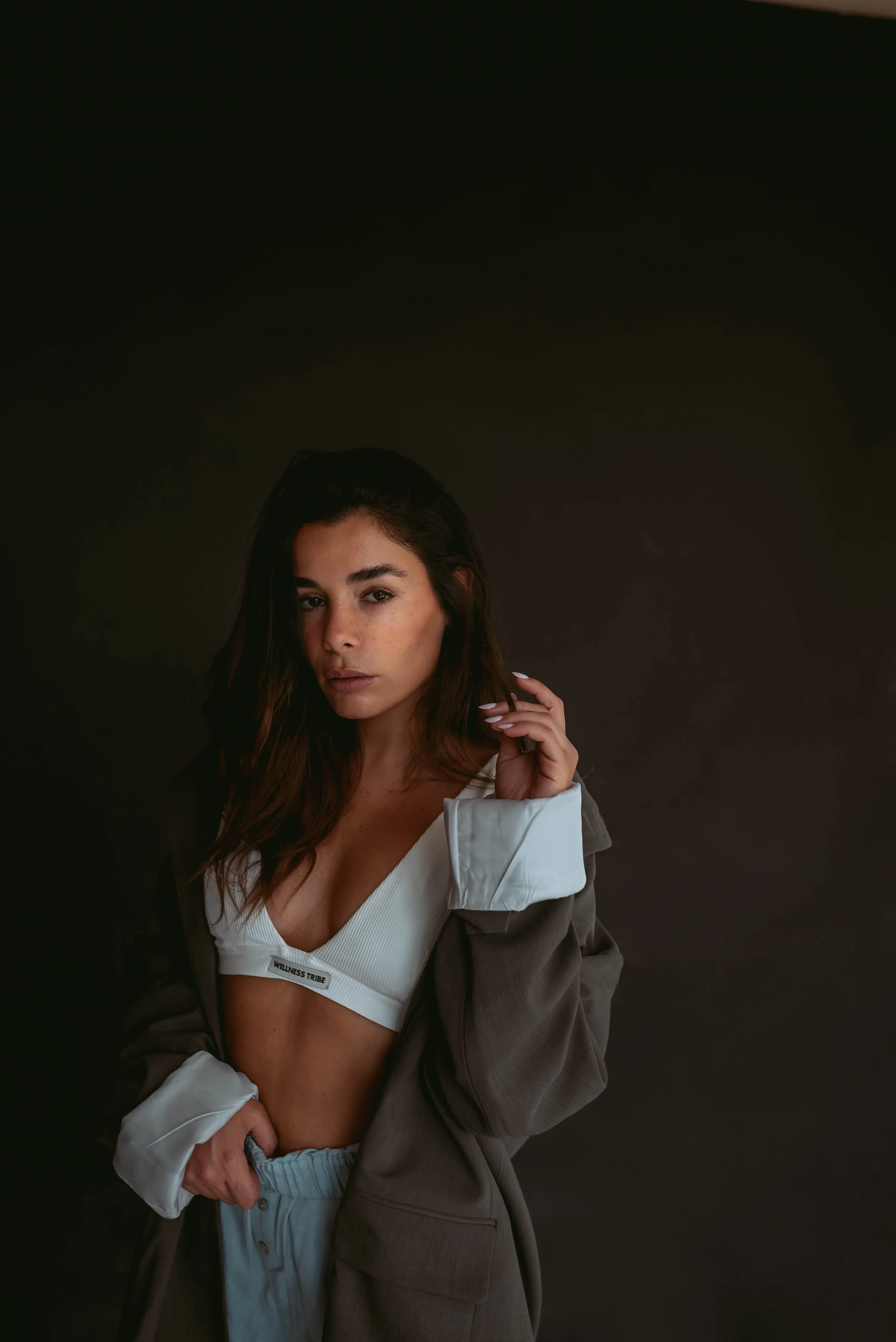 Porträt einer jungen Frau mit dunklen, gewellten Haaren, die eine off-shoulder weiße Oberteil trägt und locker eine graue Jacke hält, vor dunklem Hintergrund.
