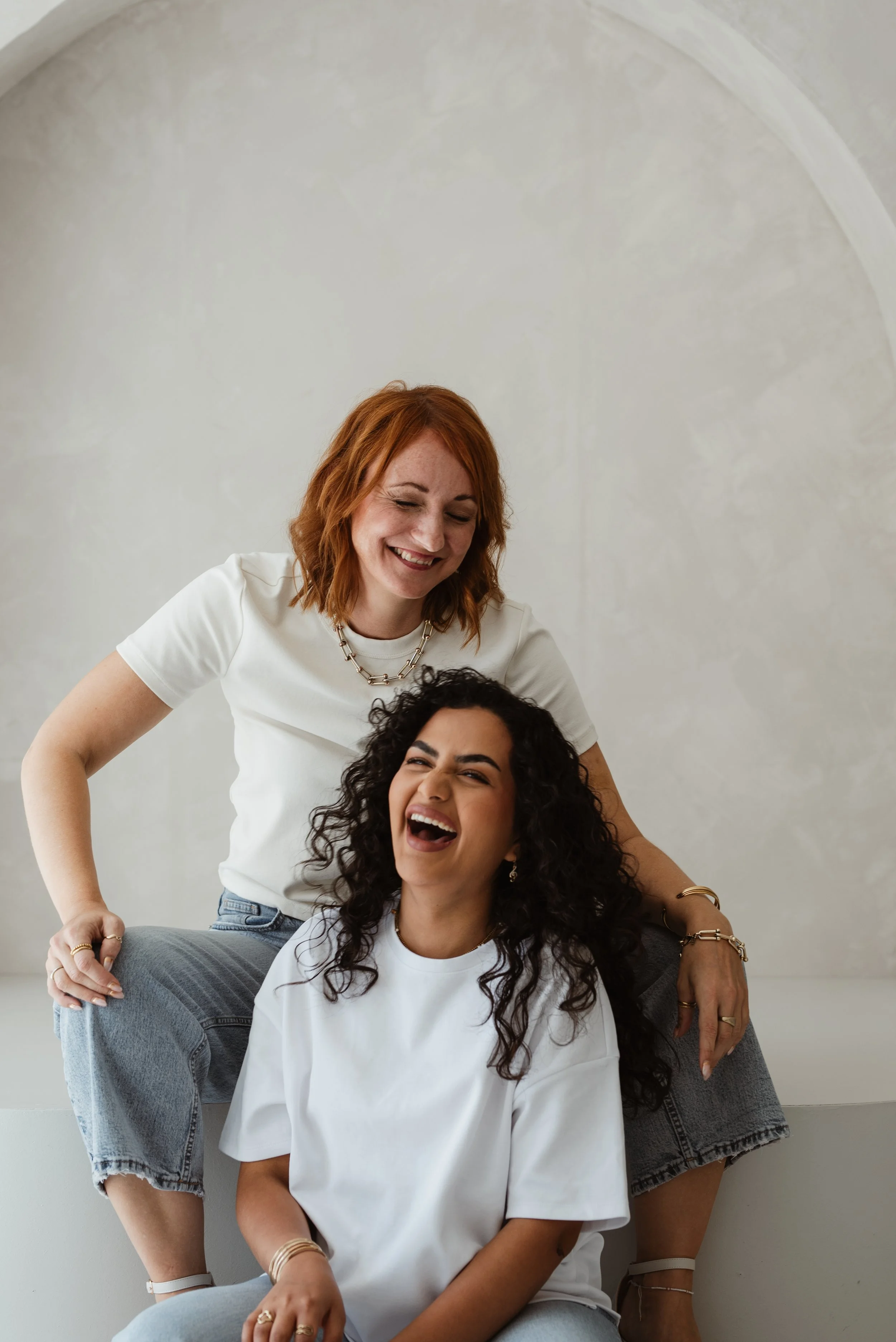 Drei Frauen lachen und posieren vor einer hellen Wand. Eine sitzt, die anderen stehen lachend um sie herum.