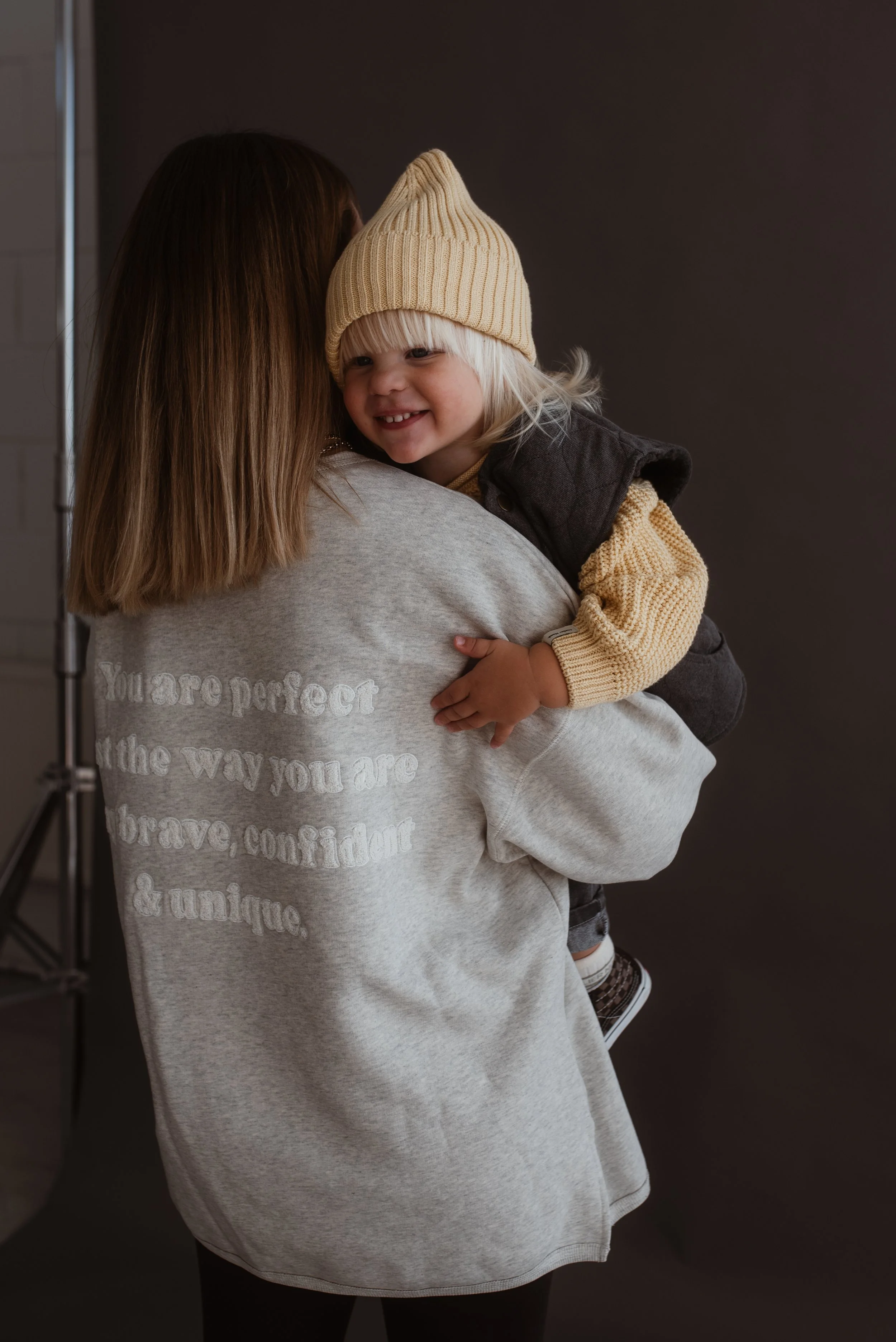 Ein Mädchen, das auf einem Arm gehalten wird, lächelt und trägt eine gelbe Strickmütze. Sie befindet sich in einer Fotostudio-Umgebung mit dunklem Hintergrund.