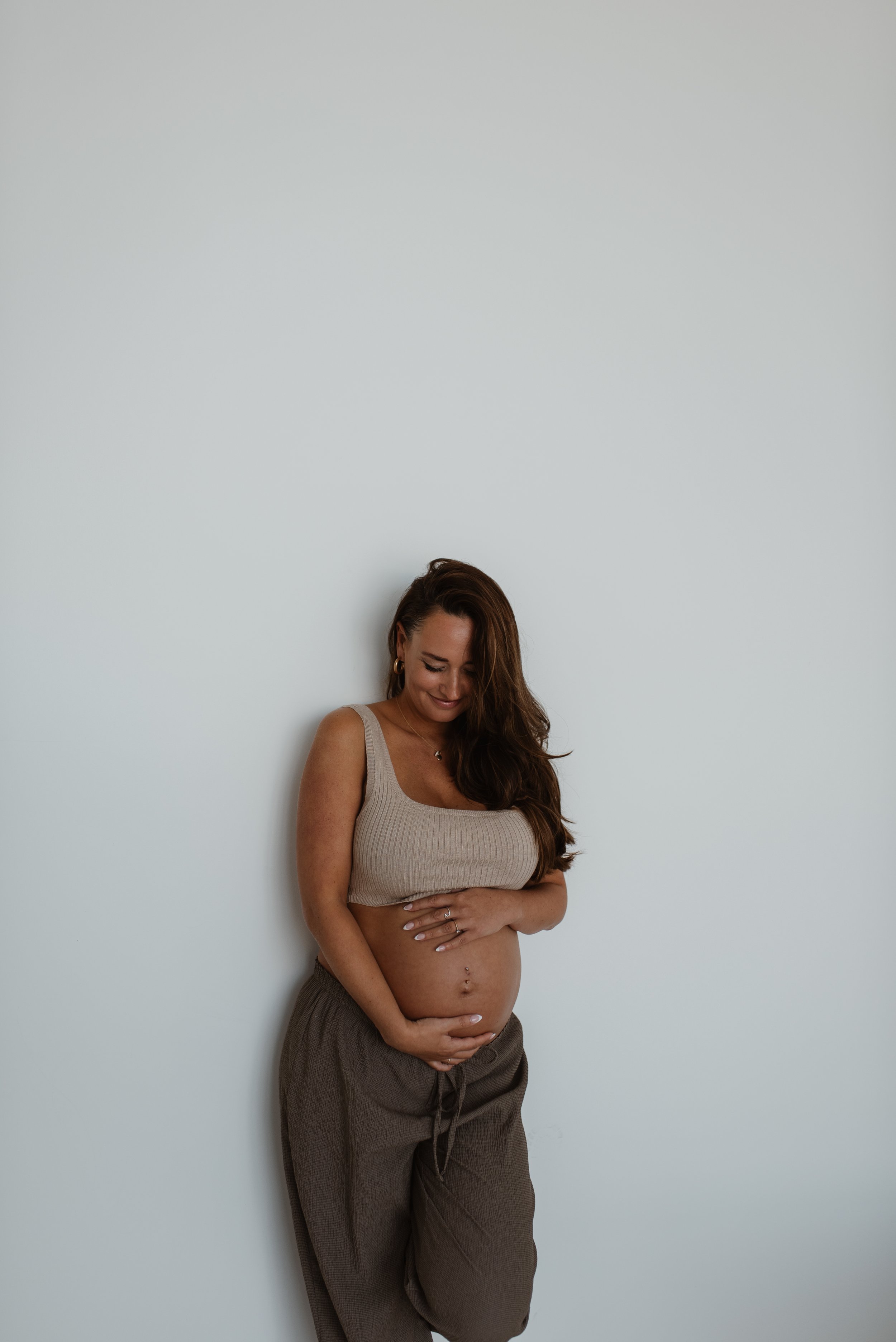 Schwangere Frau steht vor weißer Wand, trägt beiges Oberteil und braune Hose, hält den Bauch und lächelt.