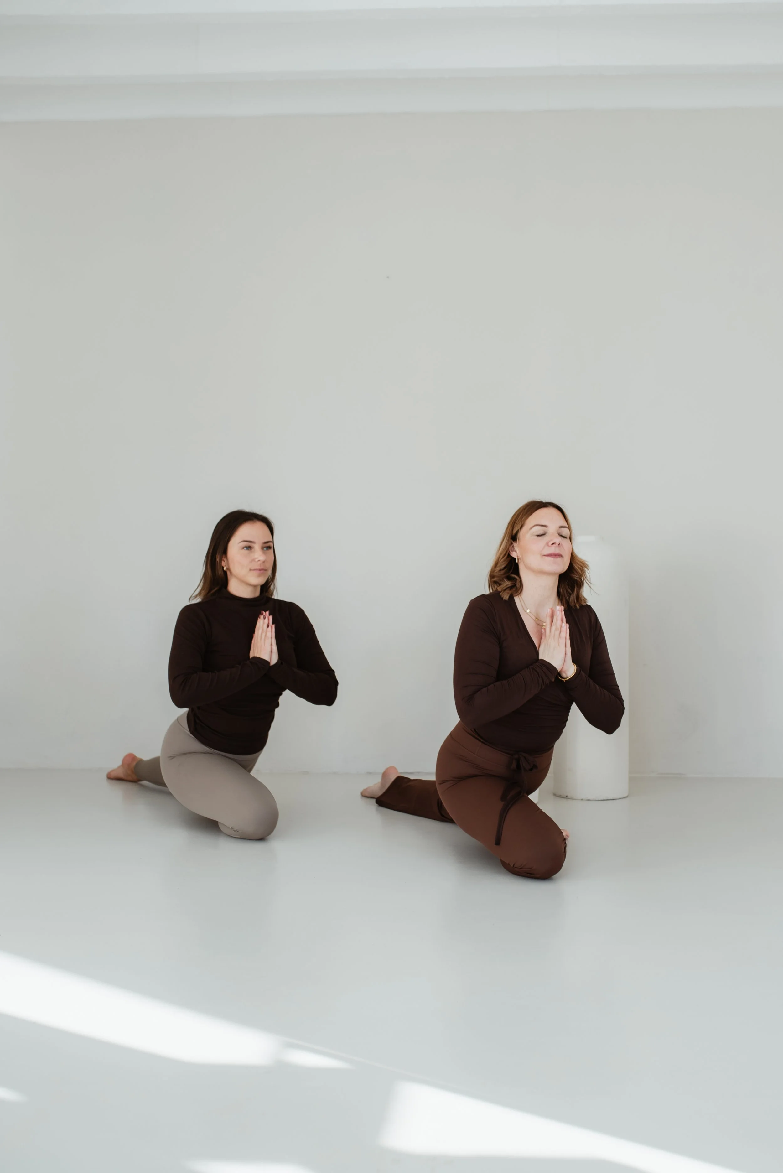 Zwei Frauen praktizieren Yoga im Schneidersitz in einem hellen Raum mit weißer Wand und Silhouette eines weißen Keramik-Topfs.