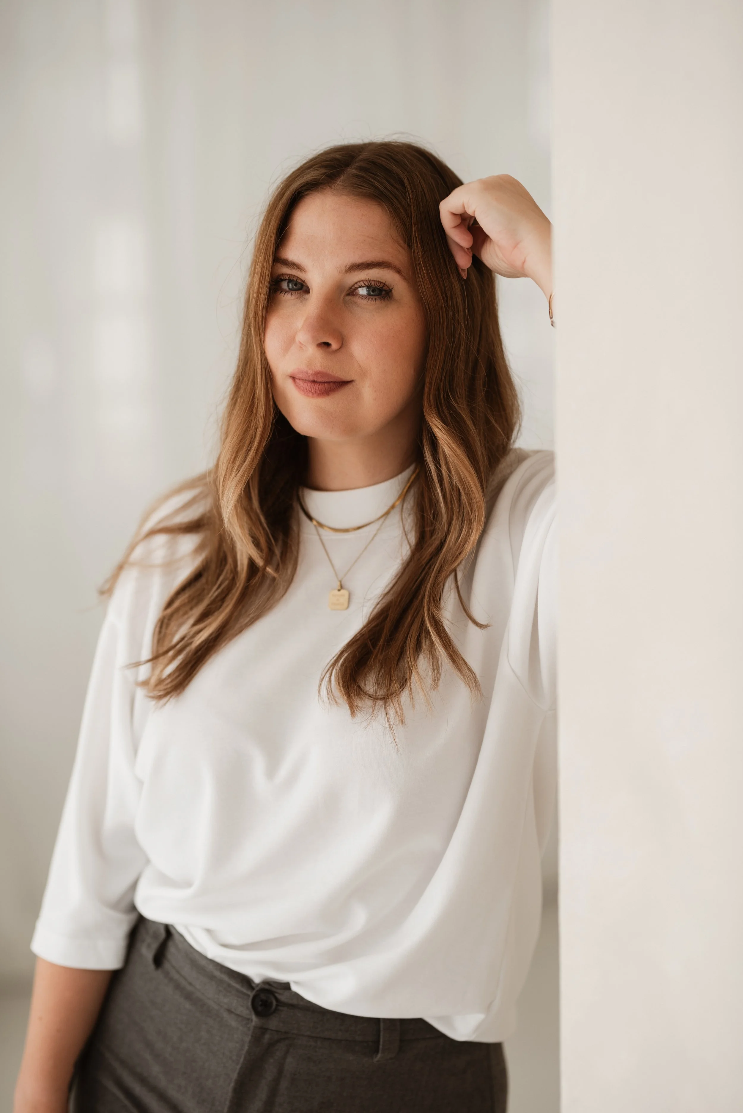 Porträt einer jungen Frau mit braunen, lockigen Haaren, die eine weiße Bluse trägt und an einer weißen Wand lehnt.