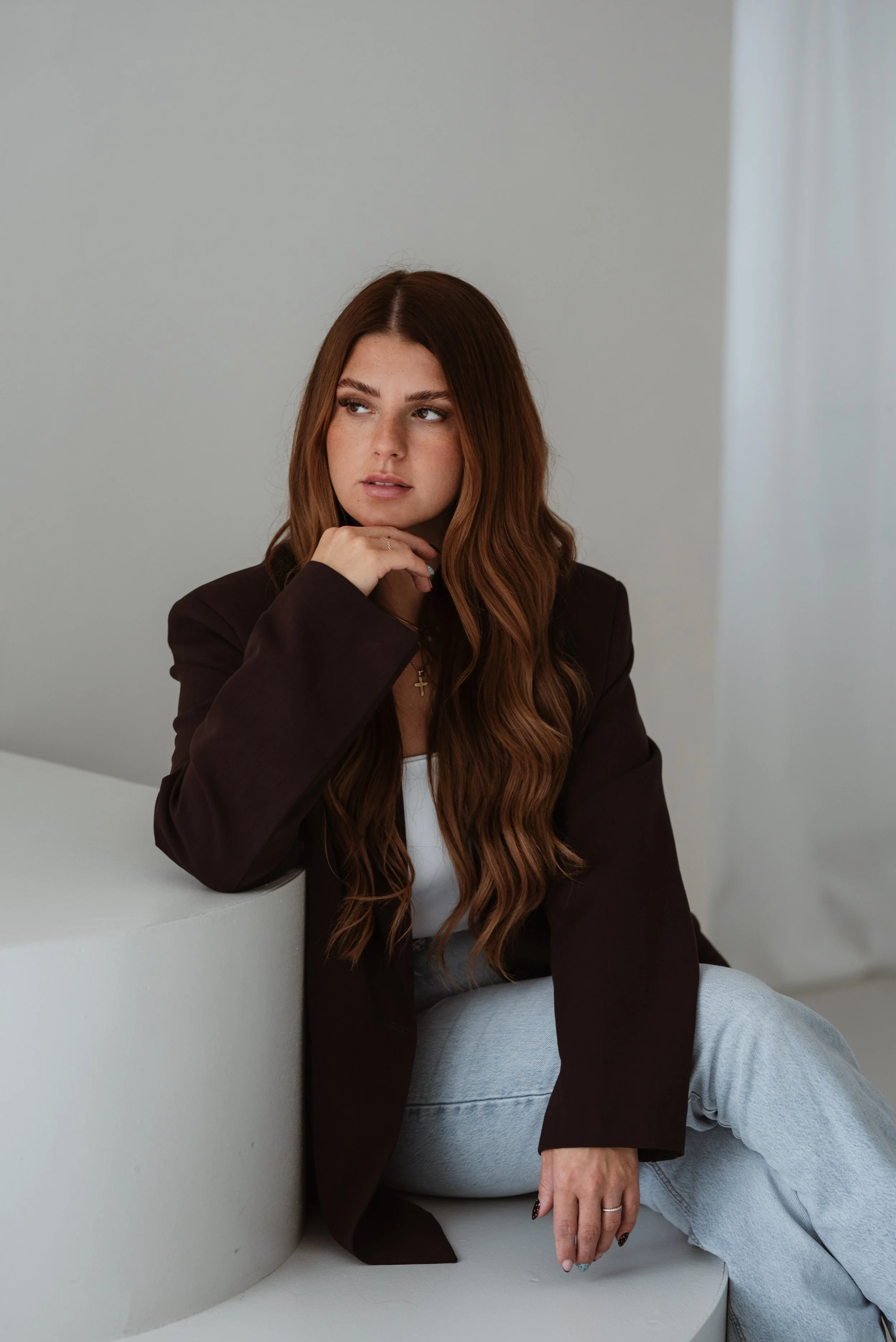 Eine junge Frau mit langen, bräunlichen Haaren sitzt auf einer weißen, gebogenen Couch vor einem hellgrauen Hintergrund. Sie trägt eine dunkle Jacke und eine weiße Top, schaut nach links und berührt mit der rechten Hand ihr Kinn.