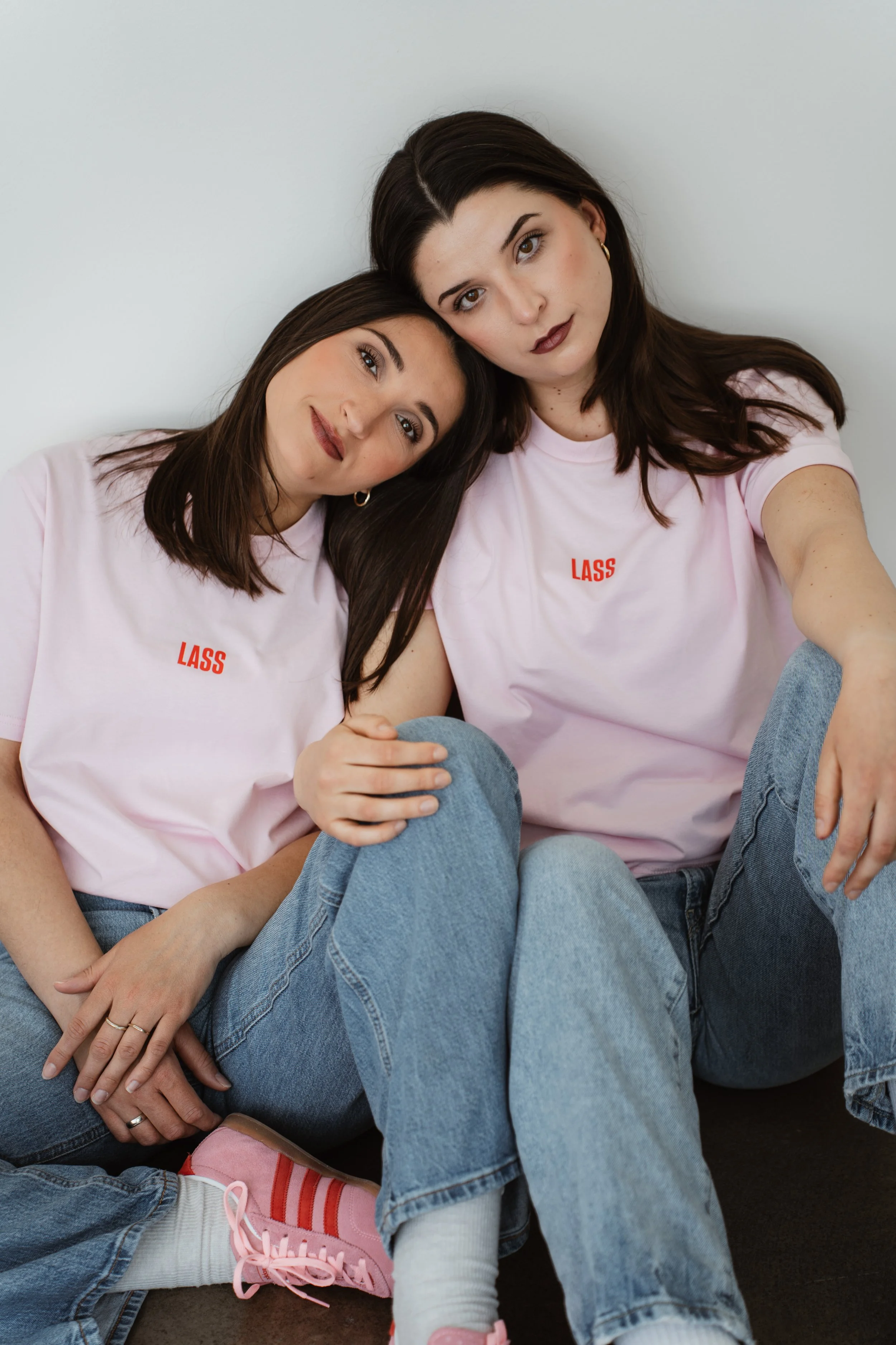 Zwei junge Frauen sitzen eng beieinander gegen eine weiße Wand, tragen helle rosa T-Shirts mit roten Buchstaben 'LASS', lächeln zärtlich und nehmen Selfies auf.