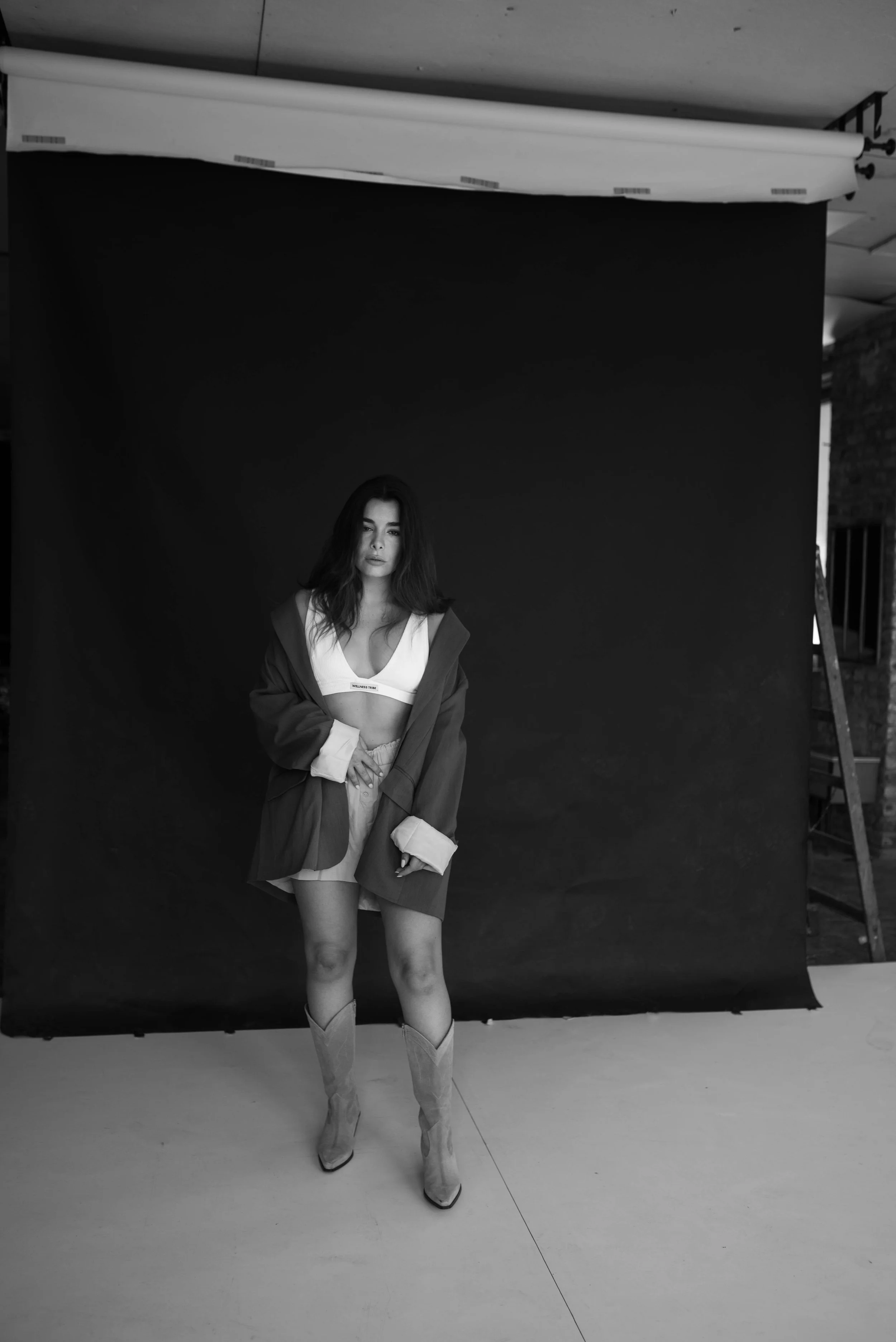 Schwarz-Weiß-Fotografie eines Models vor einem dunklen Hintergrund. Das Model trägt ein Crop-Top, eine übergroße Jacke und Stiefel.