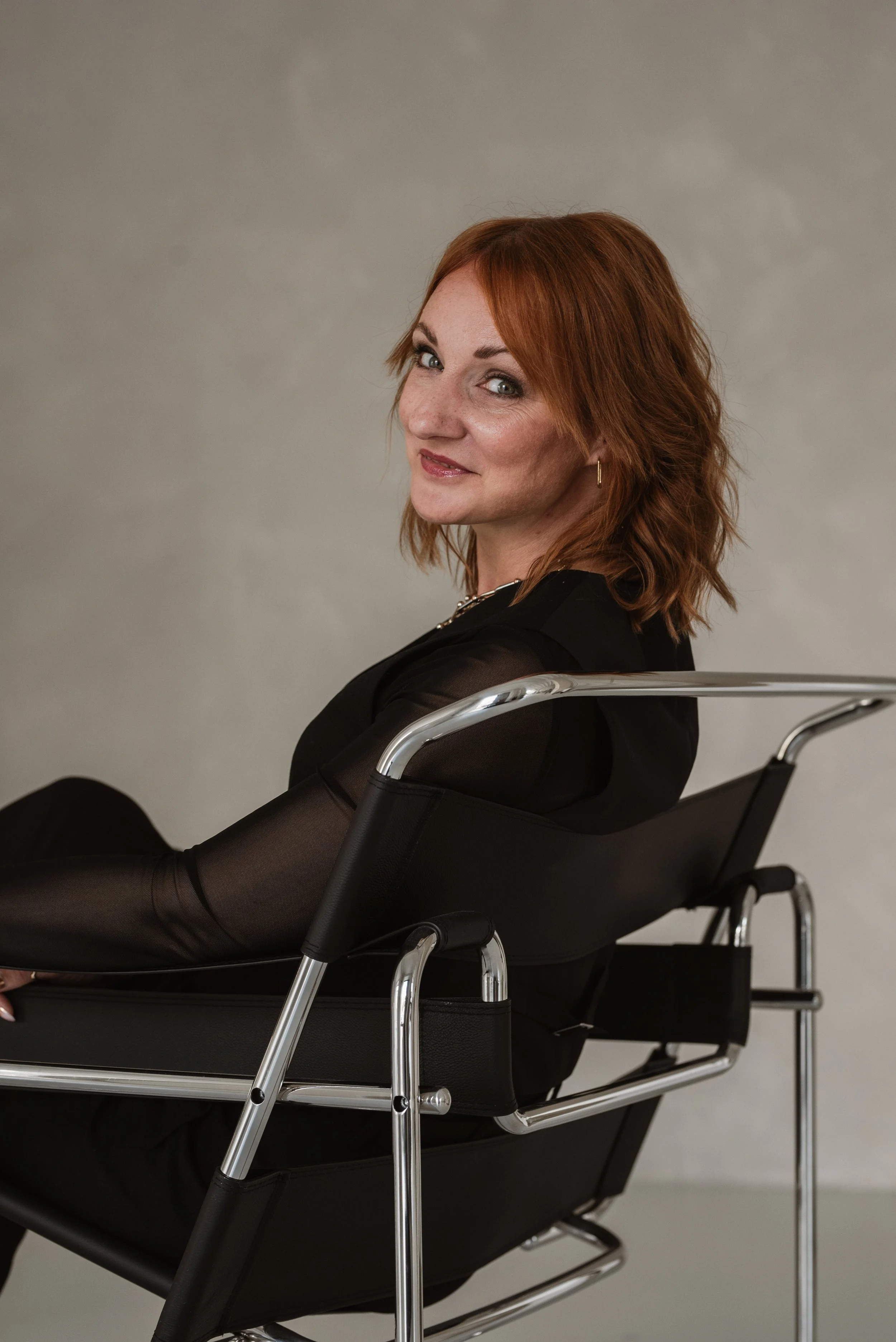 Eine Frau mit roten Haaren sitzt in einem schwarzen Rollstuhl und schaut lächelnd in die Kamera, vor einem grauen Hintergrund.