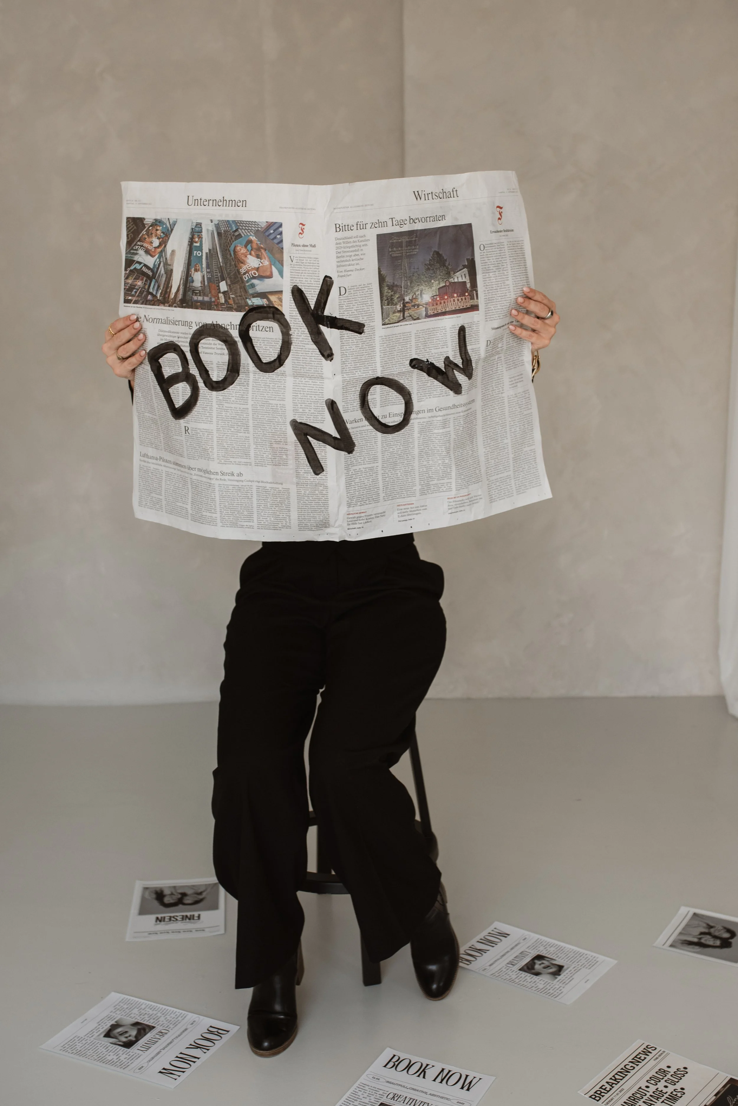 Eine Person sitzt auf einem Stuhl, hält ein zerknittertes Zeitungsblatt vor das Gesicht, auf dem in schwarzen Buchstaben "BOOK NOW" geschrieben steht, und trägt schwarze Kleidung. Es liegen mehrere Zeitungsseiten mit Überschriften auf dem Boden und a