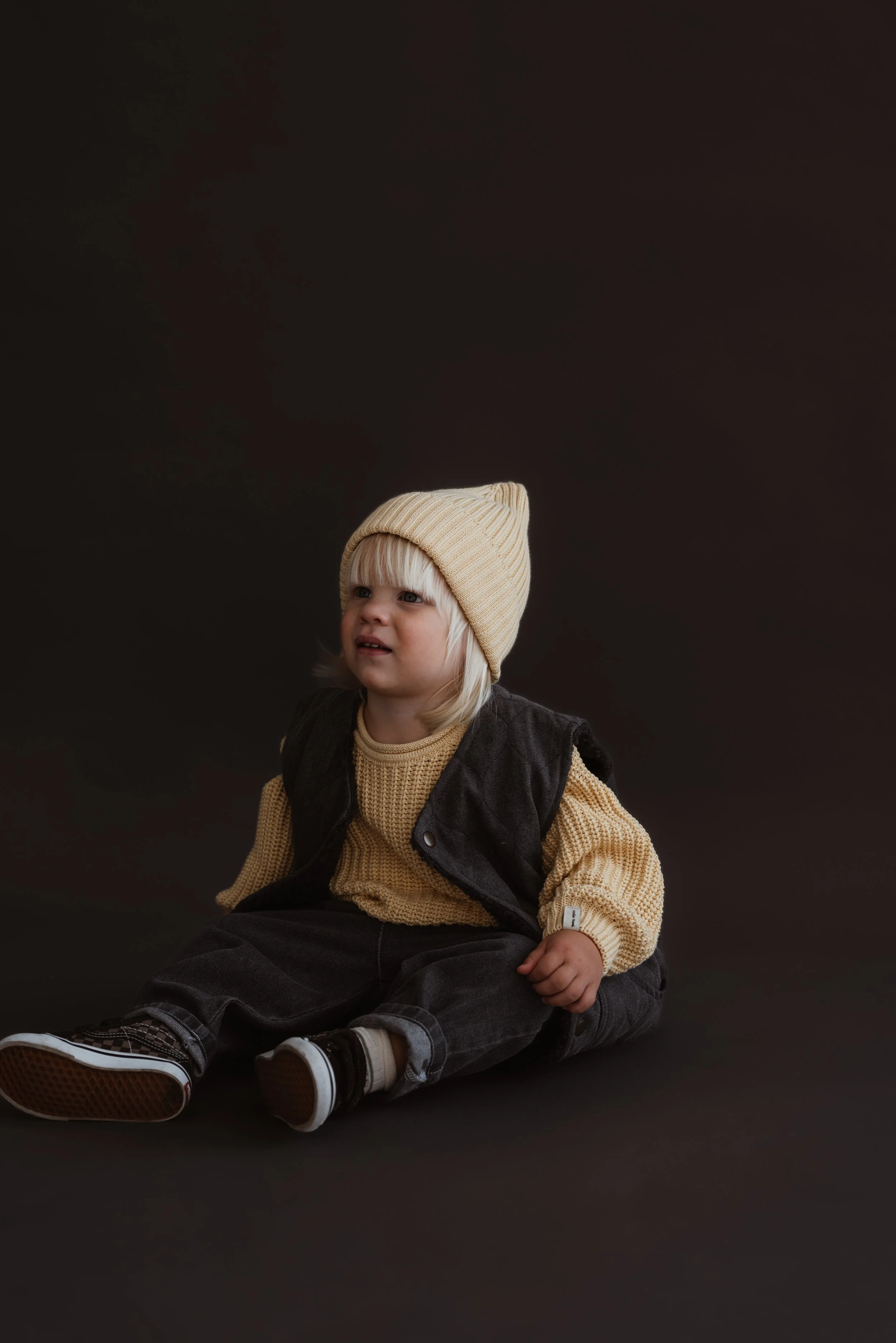 Ein kleiner Junge sitzt auf dem Boden, trägt eine beige Strickmütze, einen dunkel grau ärmellosen Pullover, eine beige Strickjacke, dunkle Hose und Sportschuhe, vor einem dunklen Hintergrund.